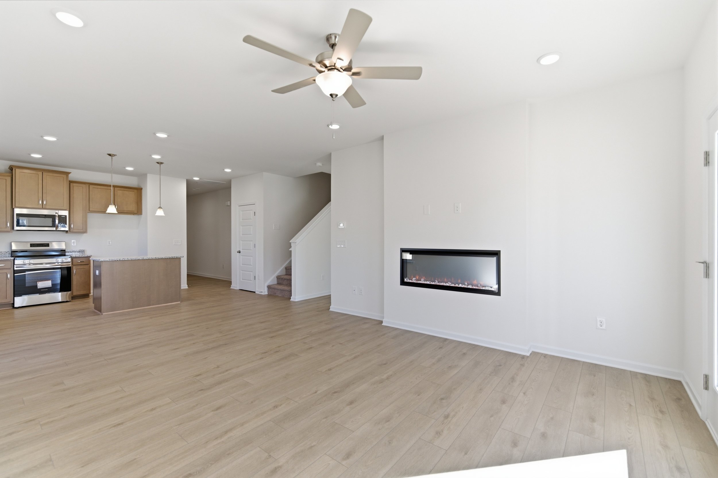 Empty living room area with wood flooring, modern electric fireplace on the wall, white walls, ceiling fan, and an open kitchen with light wood cabinets and hanging pendant lights.