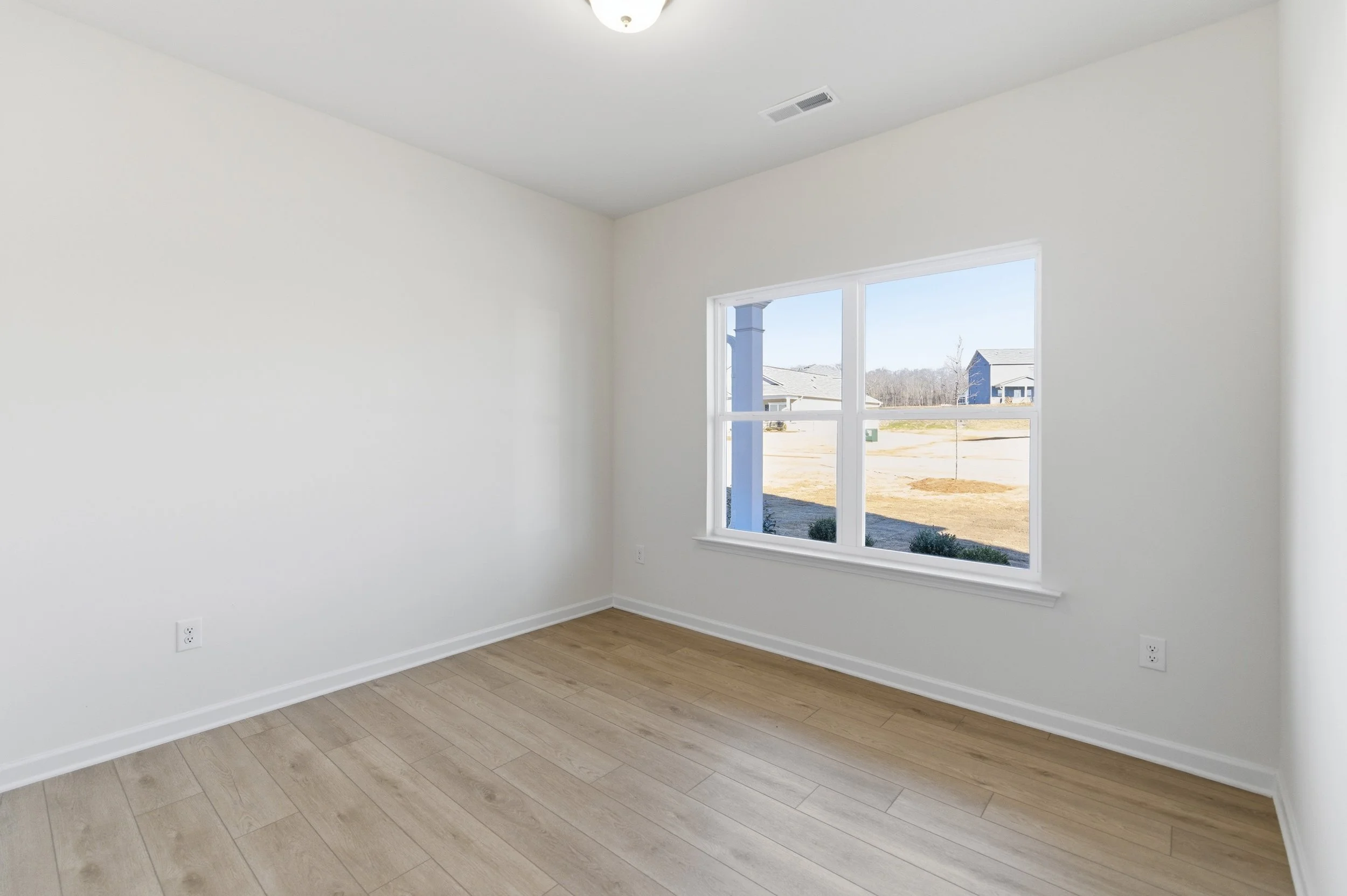 Empty room with white walls, light wood flooring, and a window showing a suburban neighborhood outside.