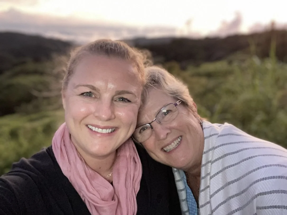 Two women smiling and leaning their heads together outdoors with a blurred background of greenery and sunset sky.