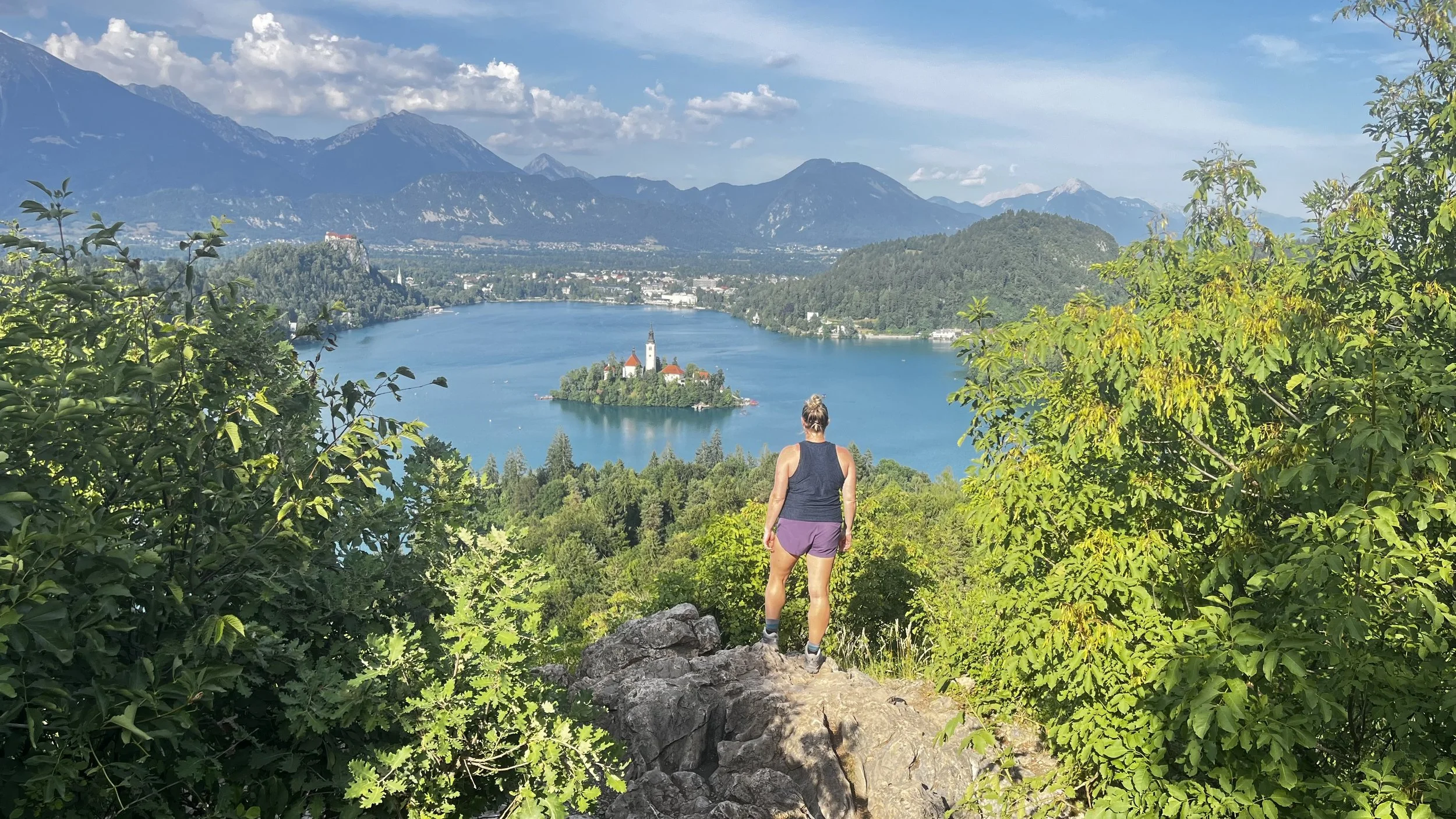 A woman standing on a rocky ledge overlooking Lake Bled with the island and church in Slovenia, surrounded by mountains and lush greenery.