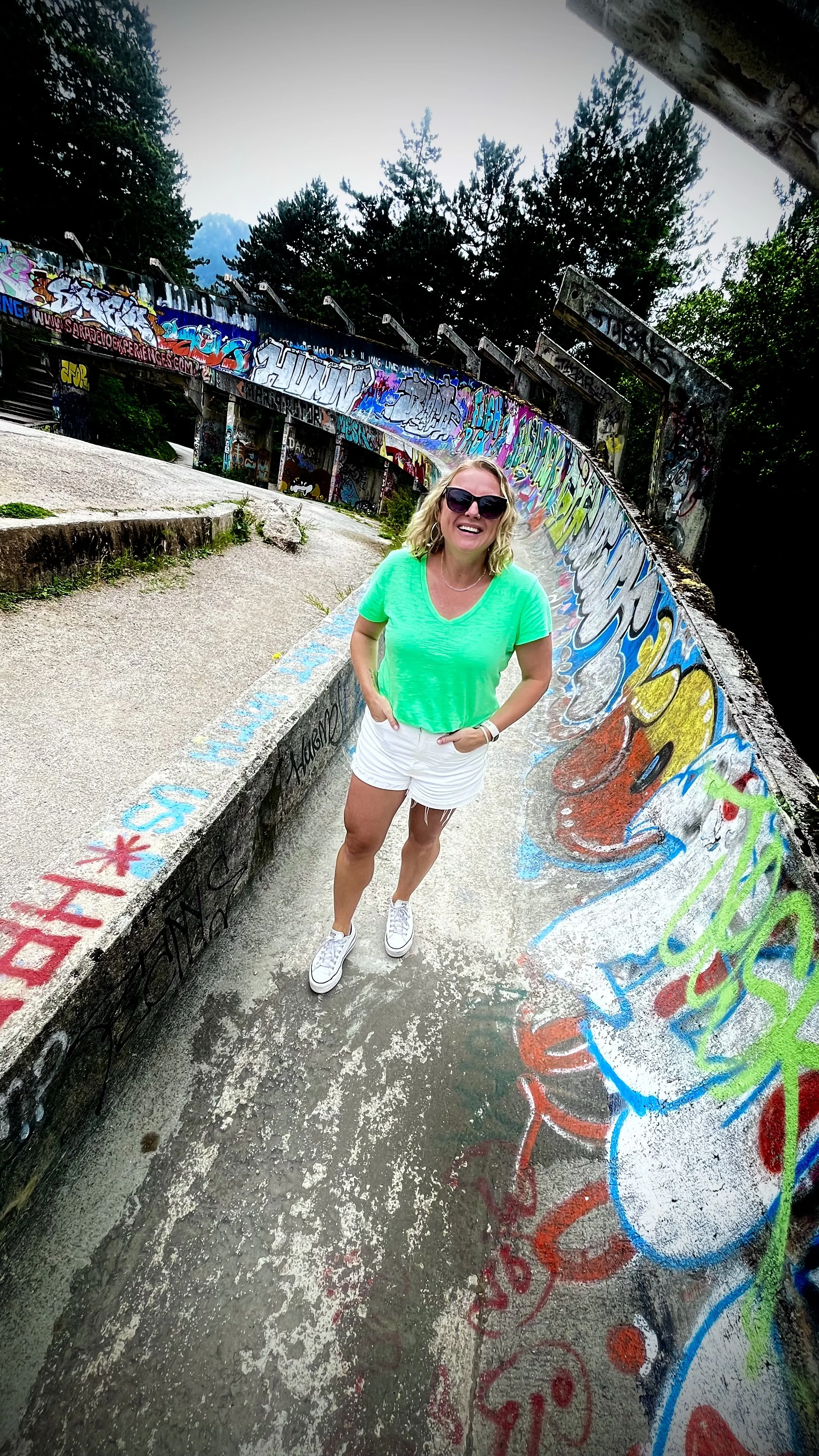 A woman wearing a bright green T-shirt and white shorts standing on a graffiti-covered concrete path, smiling and wearing sunglasses. Behind her is a curved wall painted with colorful graffiti art, with trees and a cloudy sky in the background.