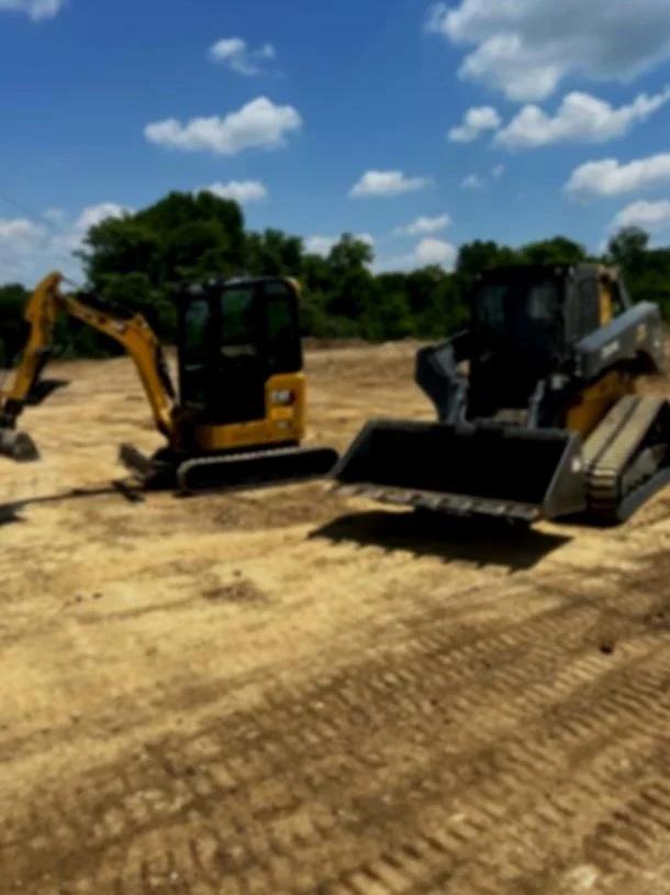 Two small yellow excavators on a construction site with bare dirt and trees in the background under a cloudy sky.