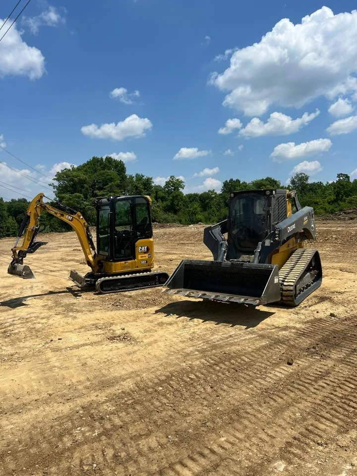 Two pieces of construction equipment, a small yellow CAT excavator and a larger black and yellow Deere compact track loader, on a dirt construction site under a blue sky with scattered white clouds.