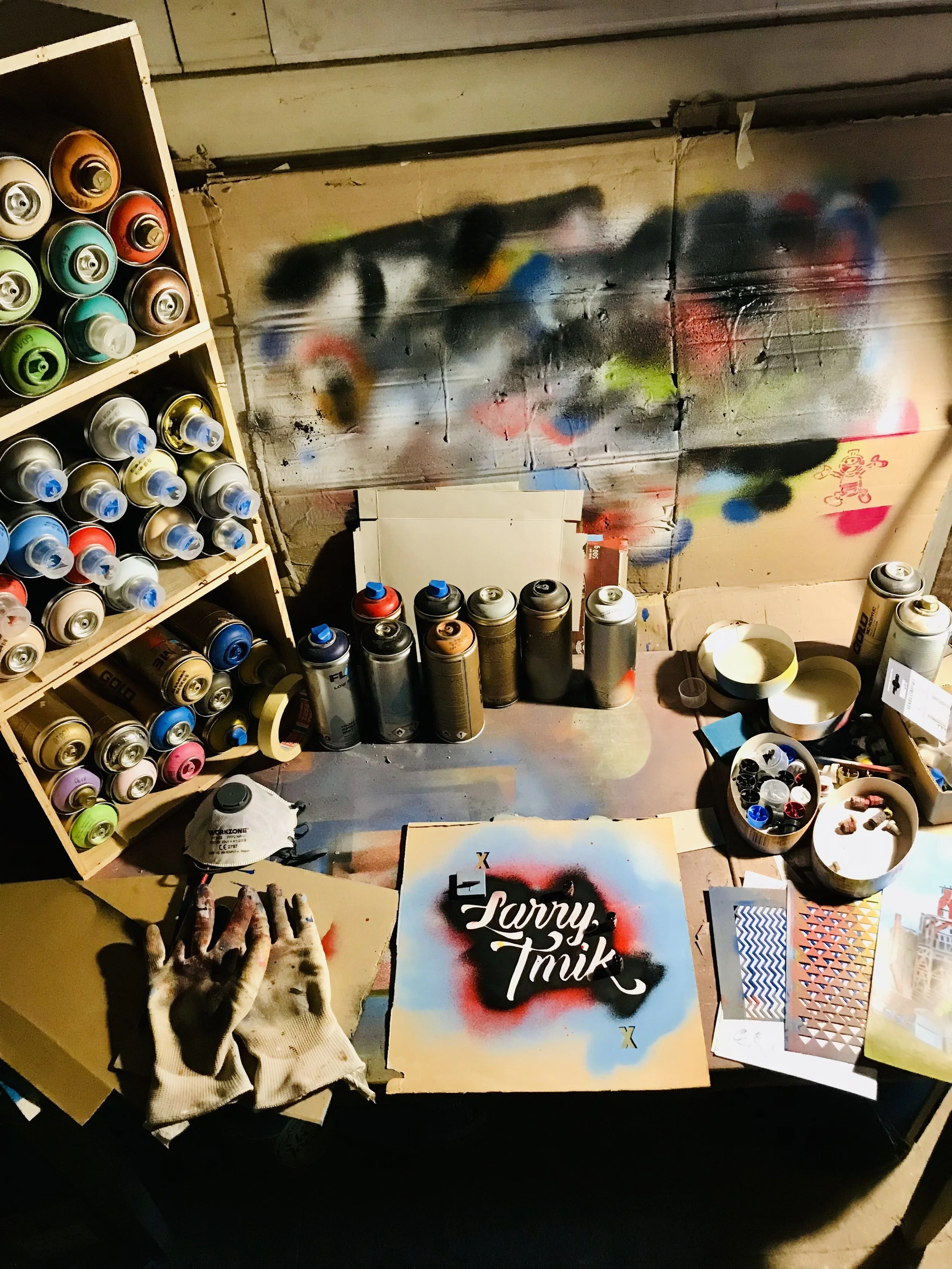 Espace de travail avec des bombes de peinture en spray, des gants, un masque, et une œuvre en spray avec le texte « Larry Tmik » sur un mur de graffiti coloré.