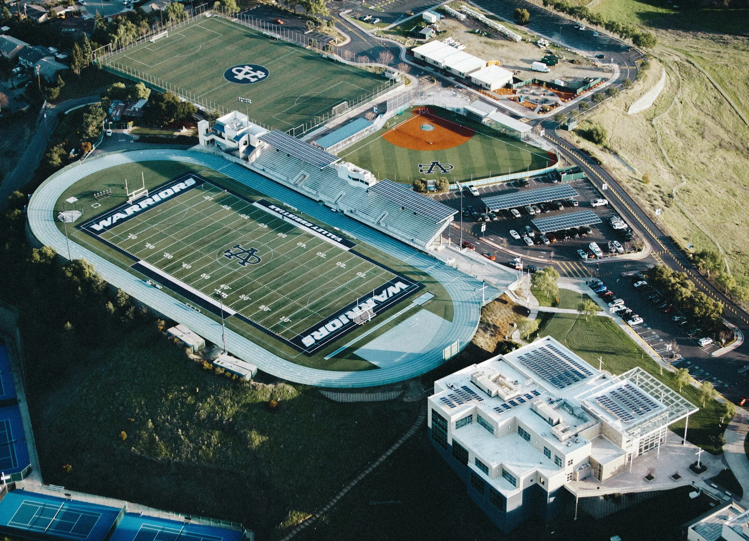 Aerial view of a sports complex with several fields including a football field marked with 'WARRIORS' and 'WARRIORS' stadium seating, a baseball field, an asphalt track, parking lots filled with cars, and surrounding pathways and buildings.