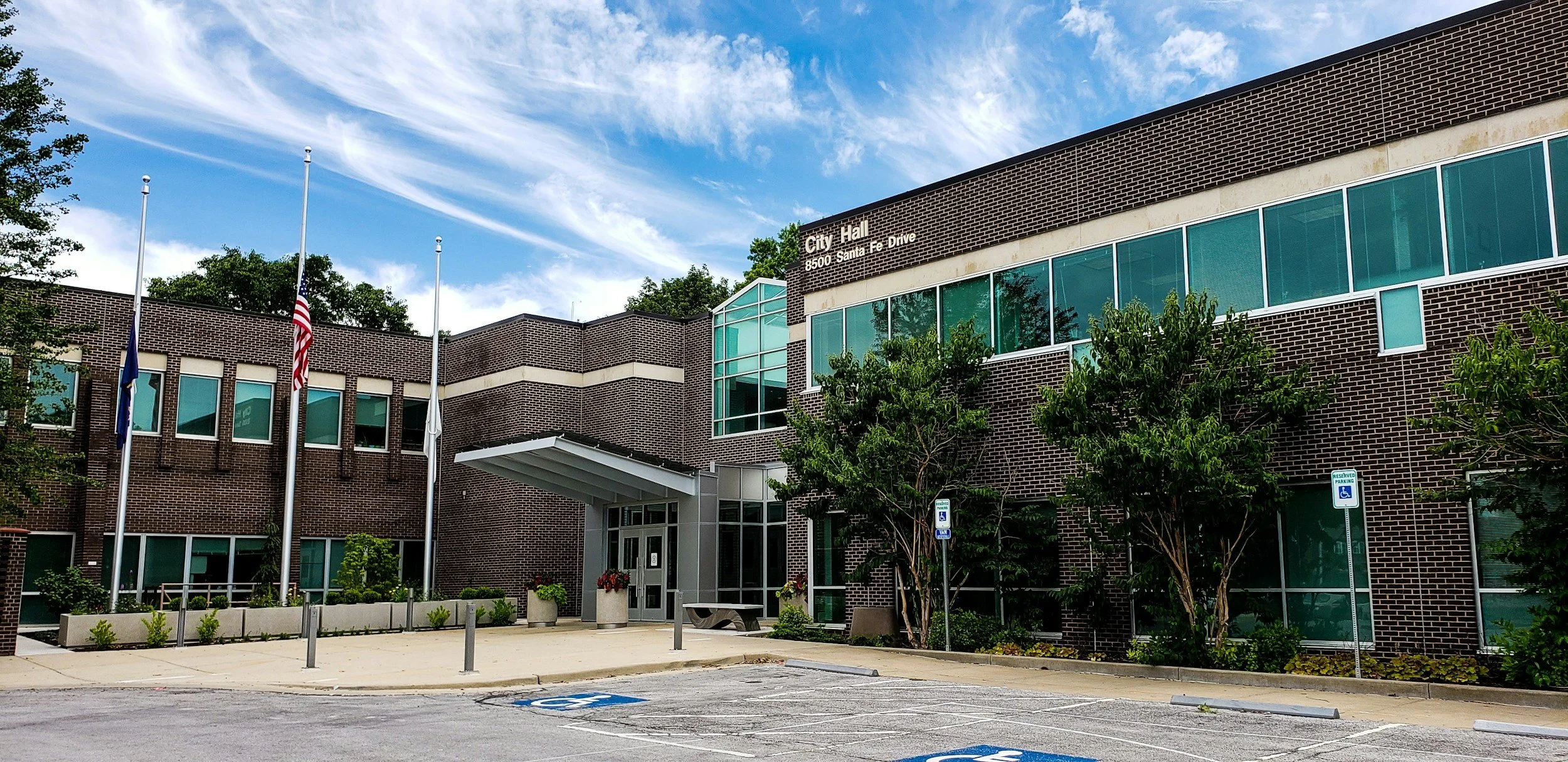 Exterior view of a city hall building made of brick with large glass windows, surrounded by trees, parking lot, flagpoles with flags, and a sign that reads 'City Hall 8500 Santa Fe Drive'.