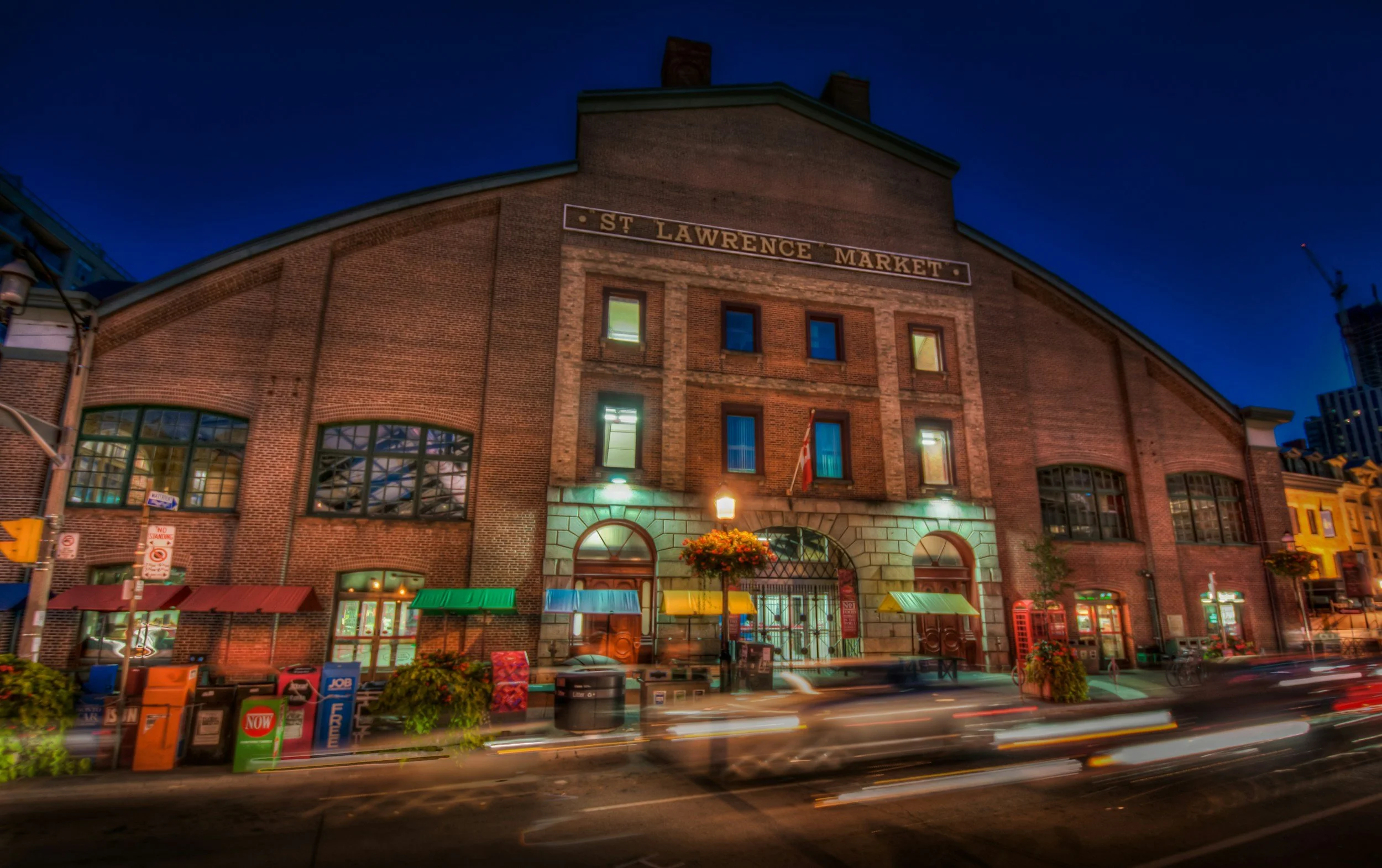 Nighttime view of St. Lawrence Market building with illuminated windows and street lights, with moving cars in front and various storefronts and newspaper boxes on the sidewalk.