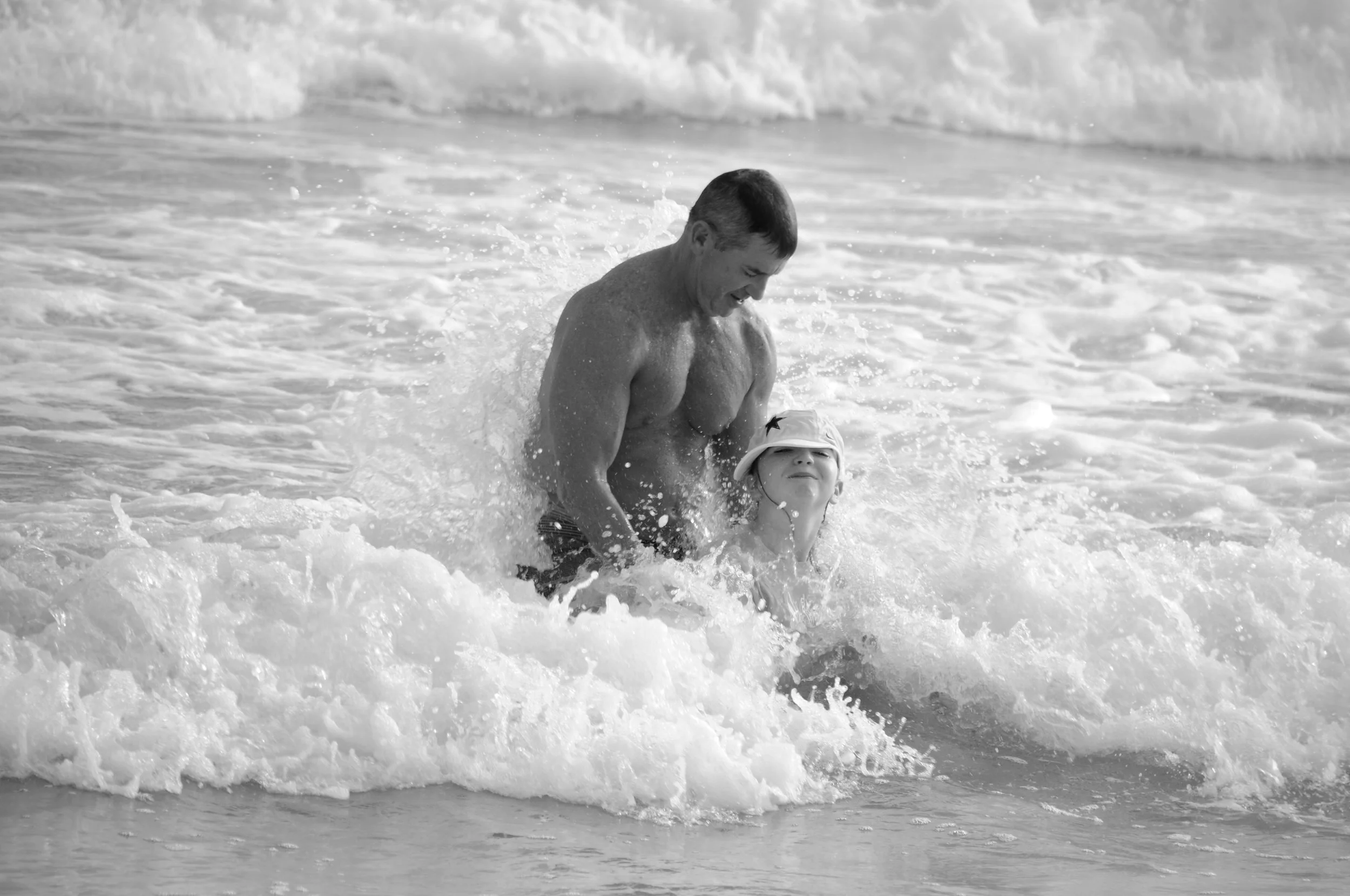 A man and a young girl playing and splashing in the water at the beach, both smiling and enjoying their time in the ocean.