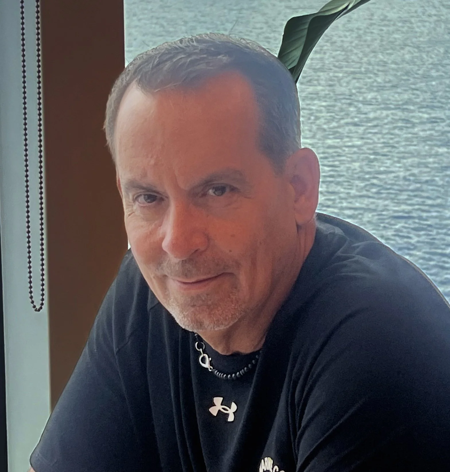 A middle-aged man with short dark hair, light facial hair, and a black Under Armour t-shirt, sitting near a window overlooking a body of water.