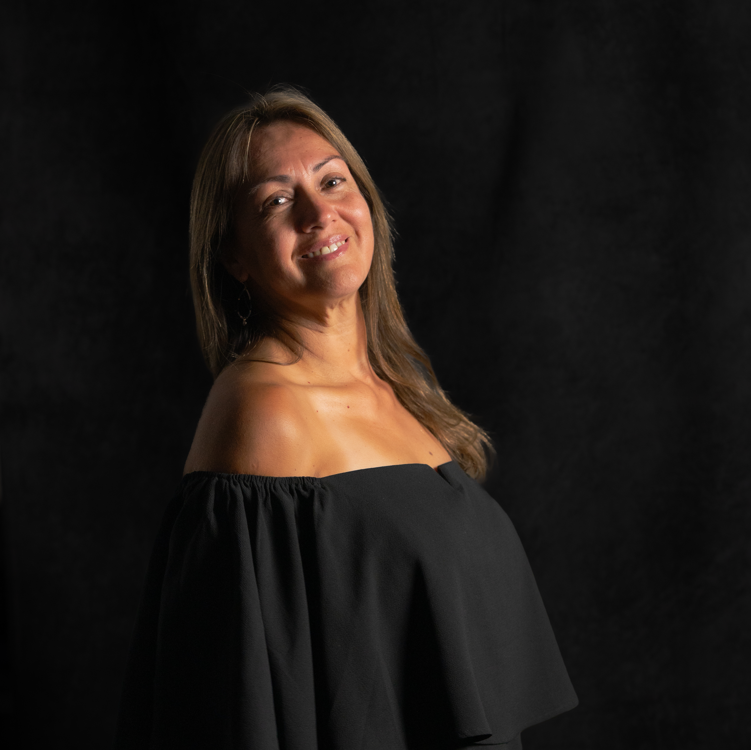 A smiling woman with long brown hair, wearing a black off-the-shoulder top, posing against a dark background.