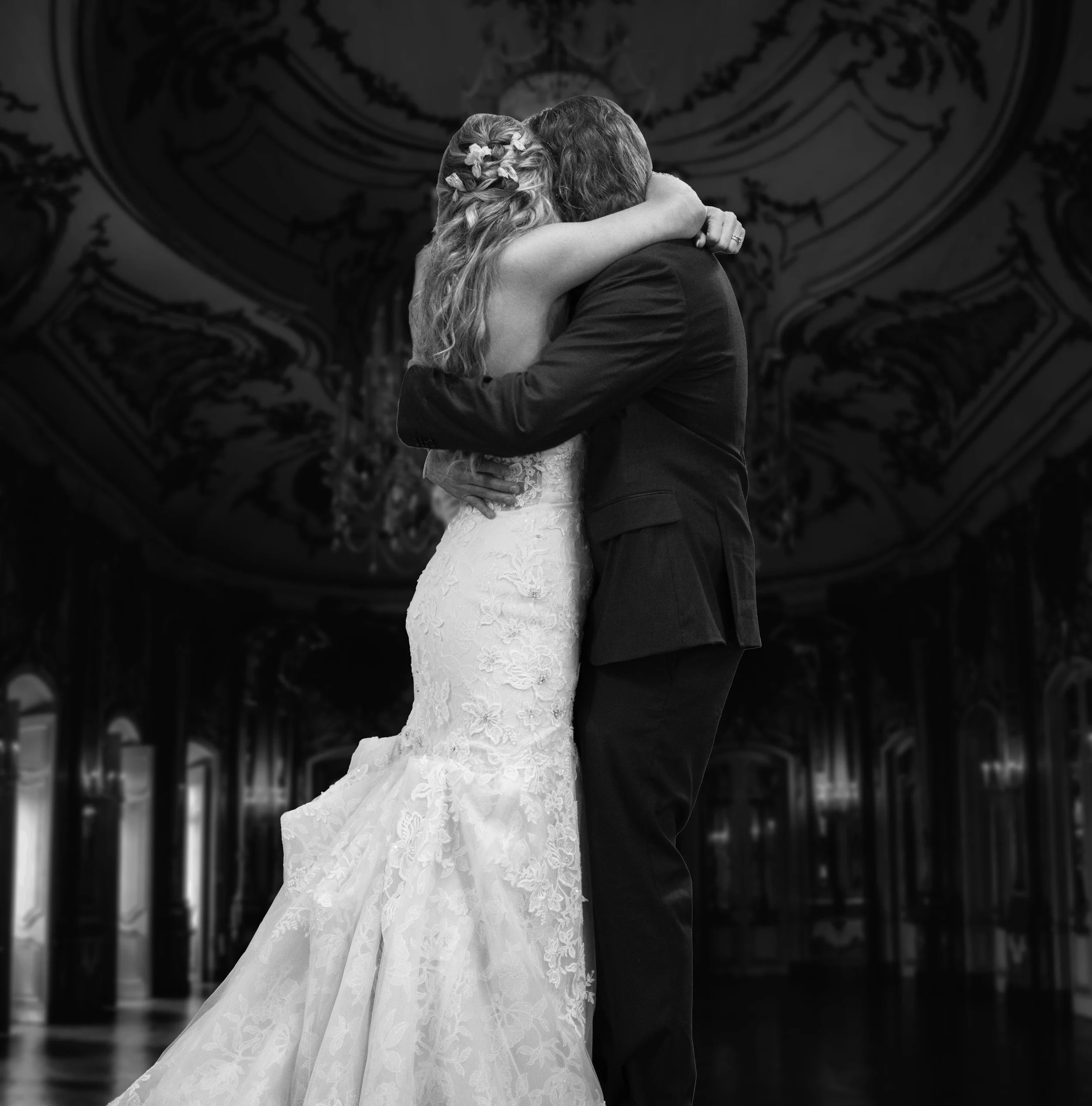 Black and white photograph of a bride and groom hugging, with the bride's arms around the groom's neck and the groom's arms around the bride's waist, in an ornate hall with a high ceiling and chandeliers.