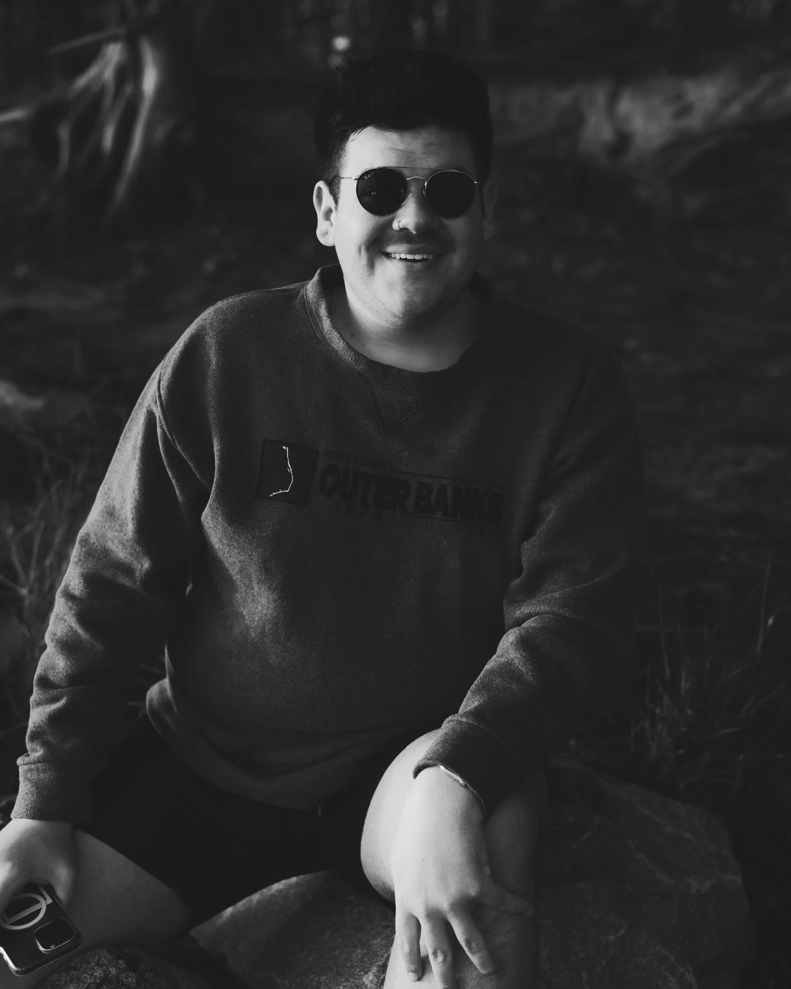 A smiling person wearing sunglasses, a sweatshirt, and shorts sitting outdoors on a rock in a black-and-white photo.