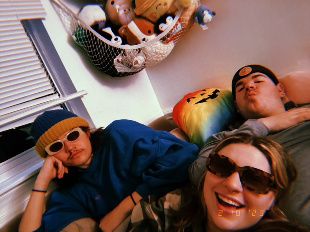Three friends relaxing on a couch, wearing sunglasses, with a box of plush toys hanging on the wall behind them, and a rainbow-colored pillow with a jack-o'-lantern on it.