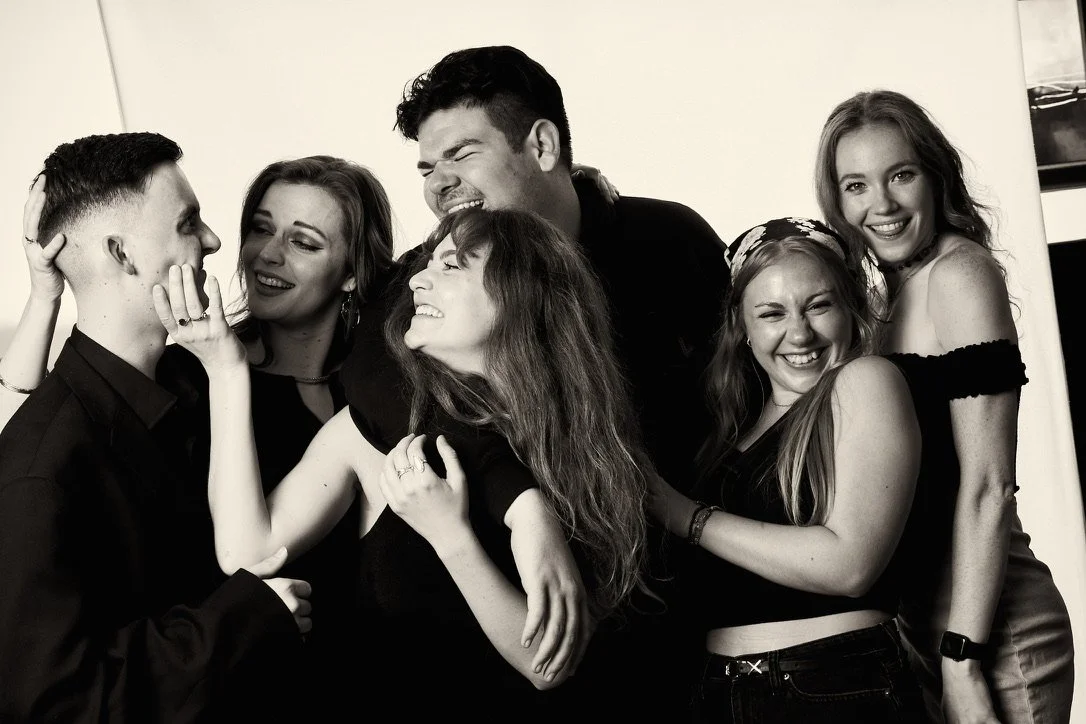 Six young adults smiling and laughing together, dressed casually, in front of a plain background.