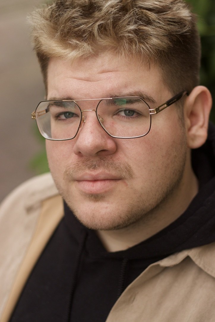 A young man with blonde hair, wearing black-rimmed glasses, a beige jacket, and a black hoodie, looking directly at the camera.