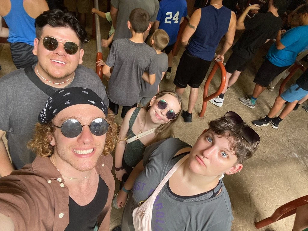 Four young people taking a selfie at a crowded indoor event, with people lining up in the background.