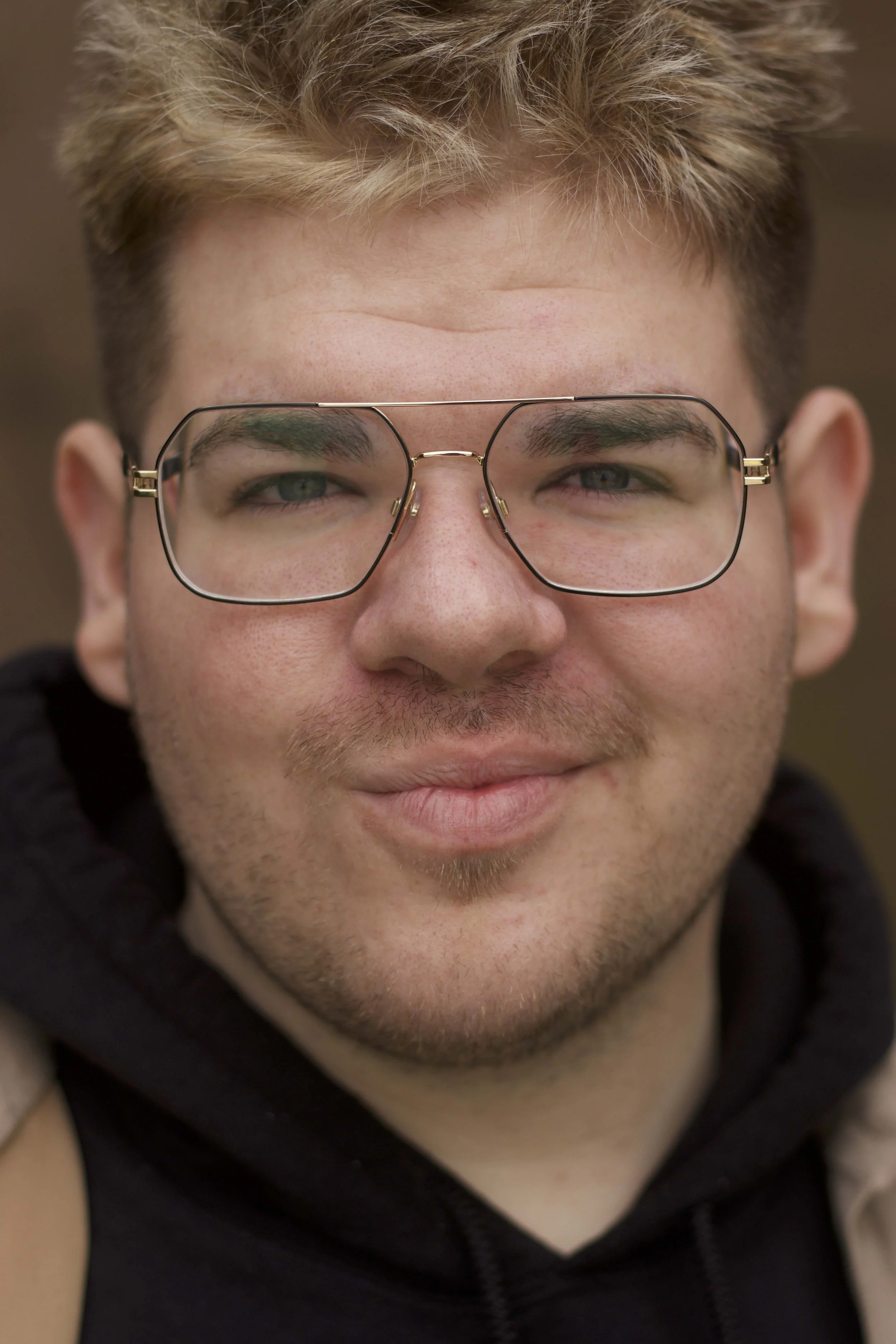 Close-up of a young man with blond hair, glasses, light skin, and a slight smile, wearing a black hoodie.
