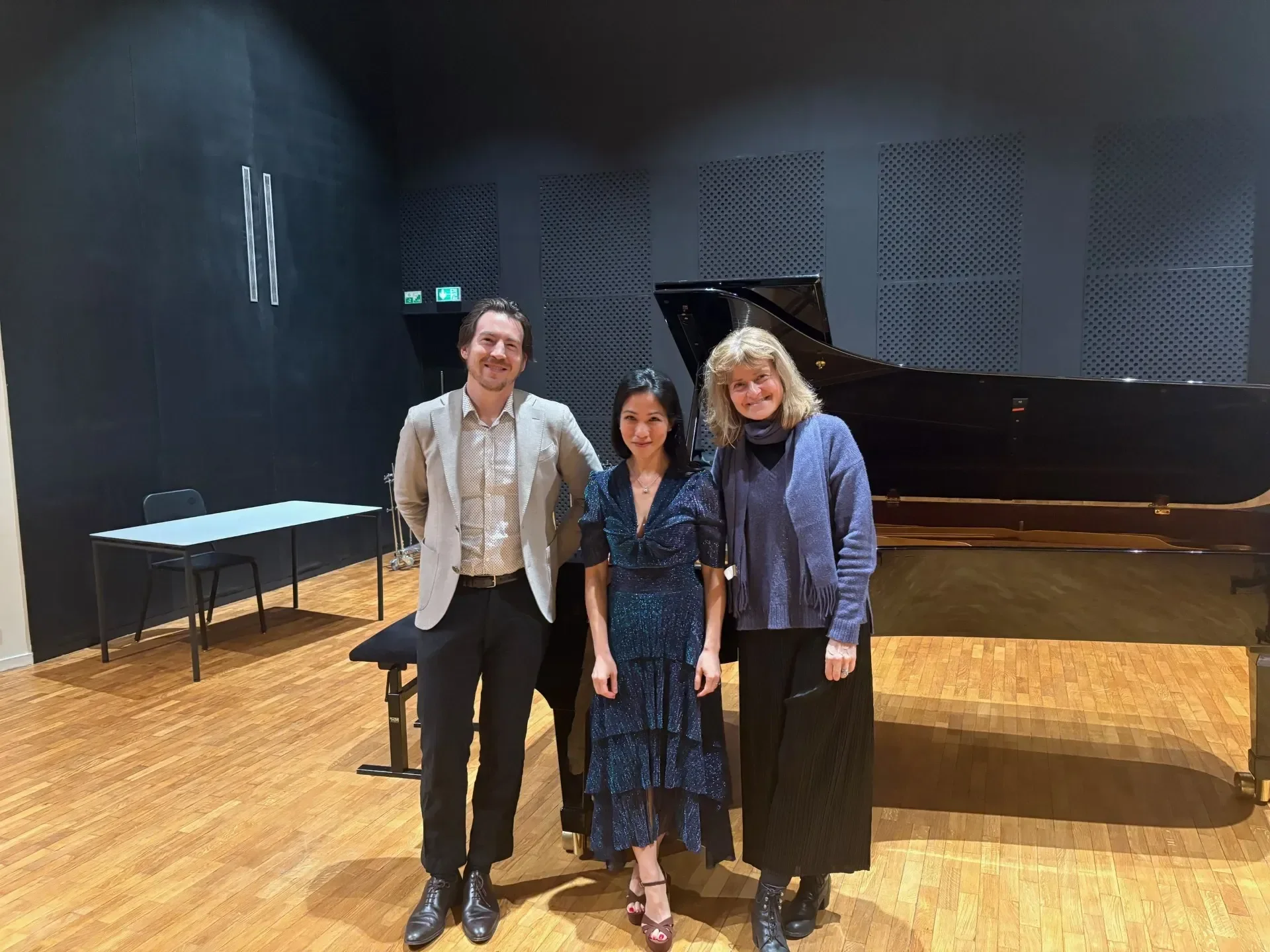 Three people standing in front of a black grand piano in a concert hall with dark walls, wooden floor, a table, and chairs in the background.