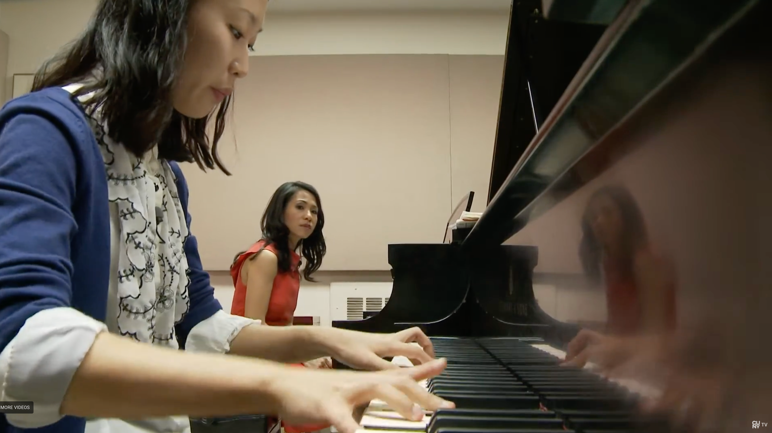Two women are playing a grand piano together, one is focused on playing, and the other is sitting in the background watching.