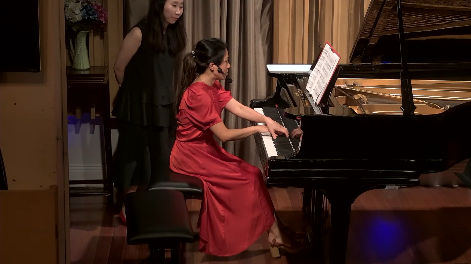 A woman in a red dress playing a grand piano with a woman in black standing nearby watching.