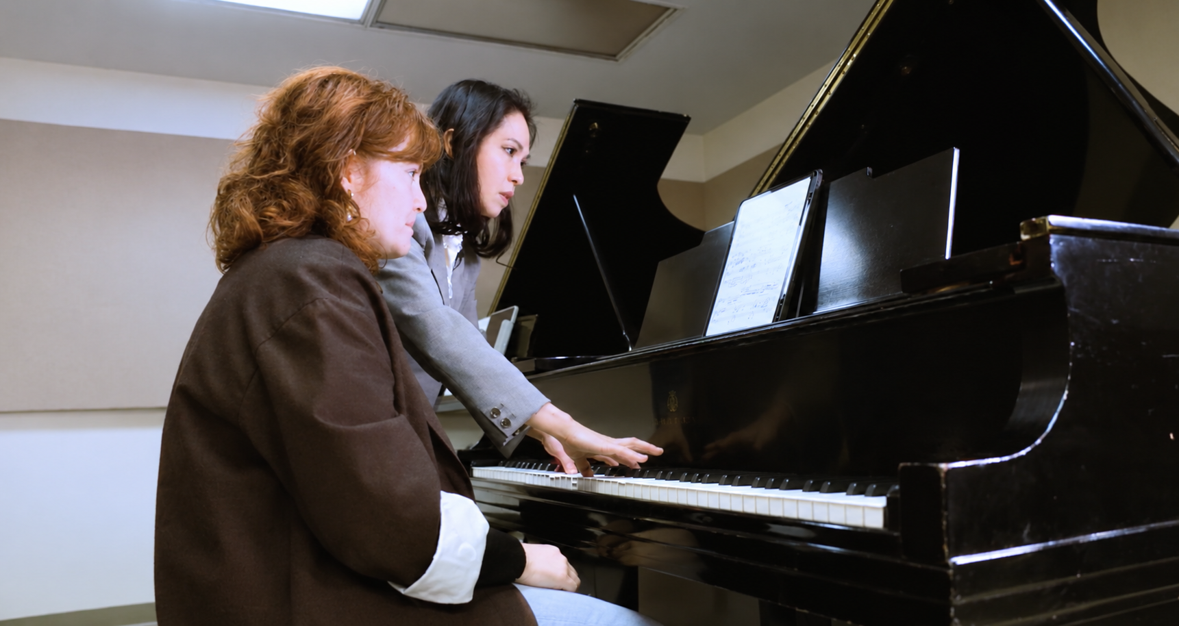Two women playing piano together, with sheet music in front of them, in a music room or studio.