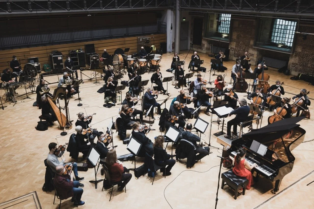 Orchestra performing in a concert hall, with musicians playing string, wind, and percussion instruments, and a pianist at a grand piano.