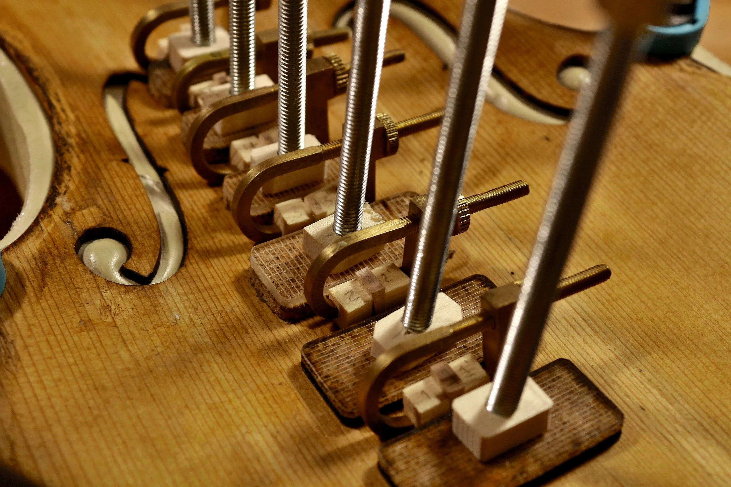 Nahaufnahme eines Holzgeigenhalses im Herstellungsprozess, mit mehreren Klemmen, die verschiedene Komponenten des Instruments fixieren.