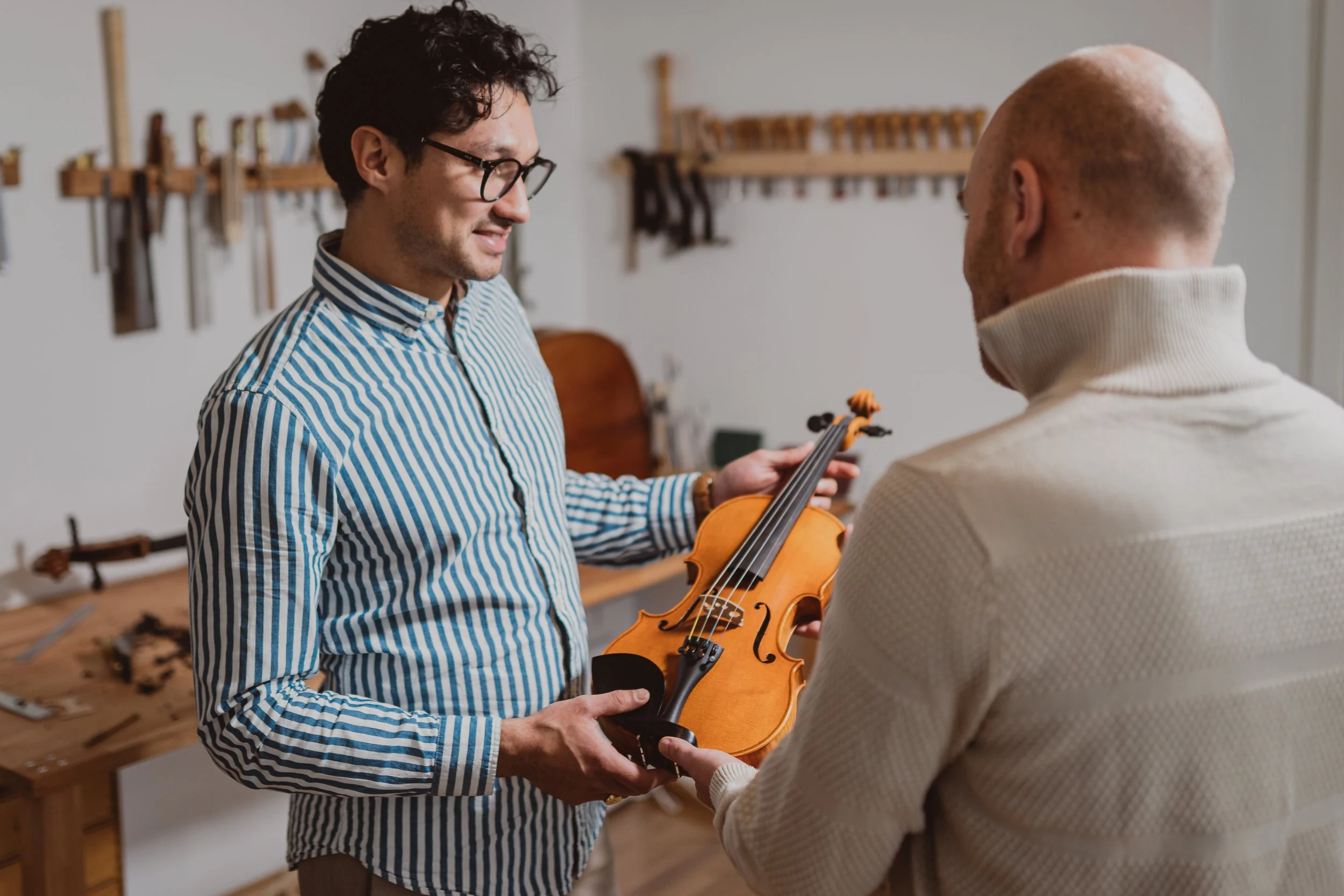 Zwei Männer in einem Raum, einer zeigt dem anderen eine Geige, im Hintergrund Werkzeuge an der Wand.