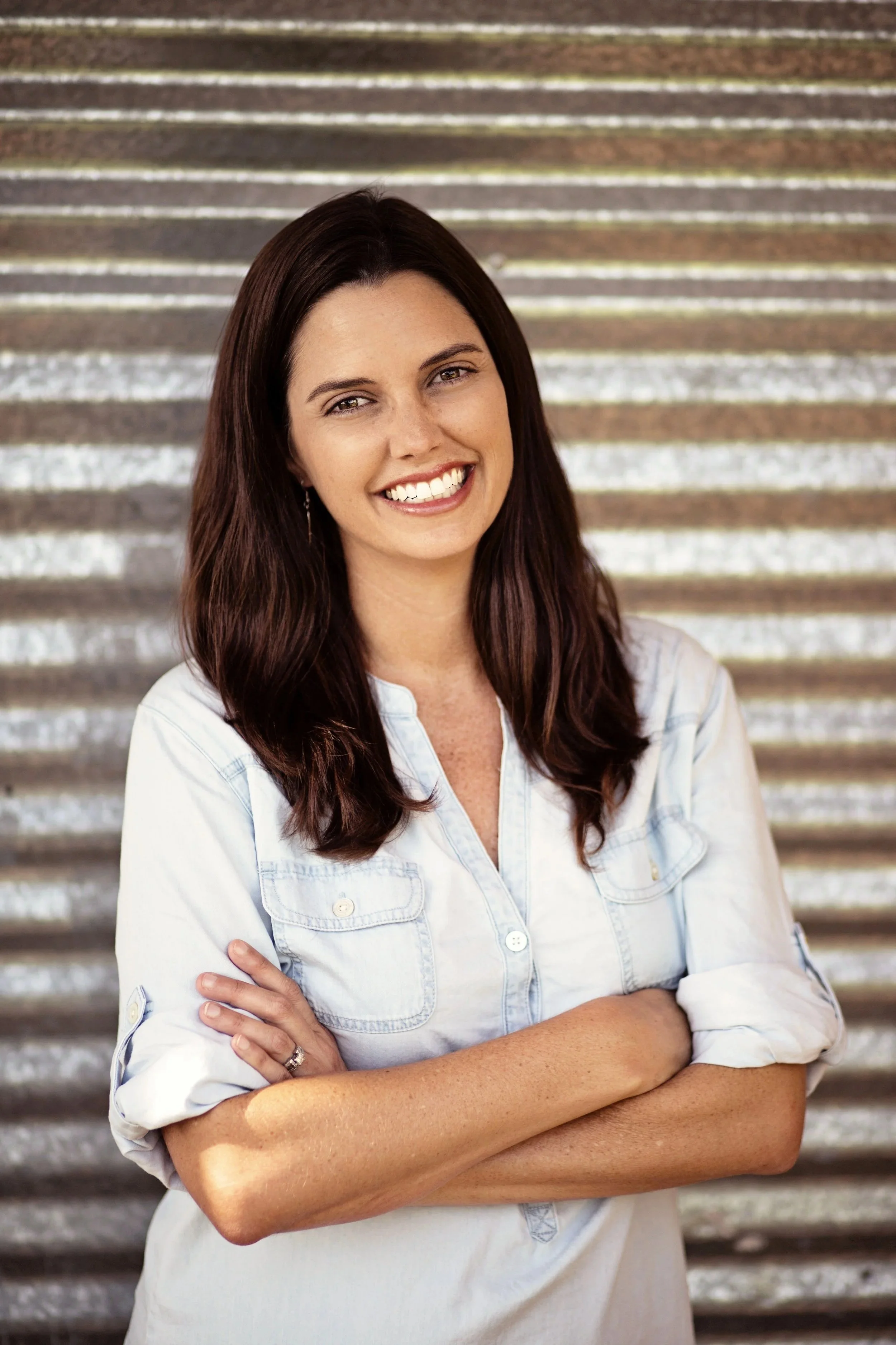 Young woman with long dark brown hair, smiling with teeth visible, arms crossed, wearing a light denim shirt, standing outdoors in front of metal stairs.