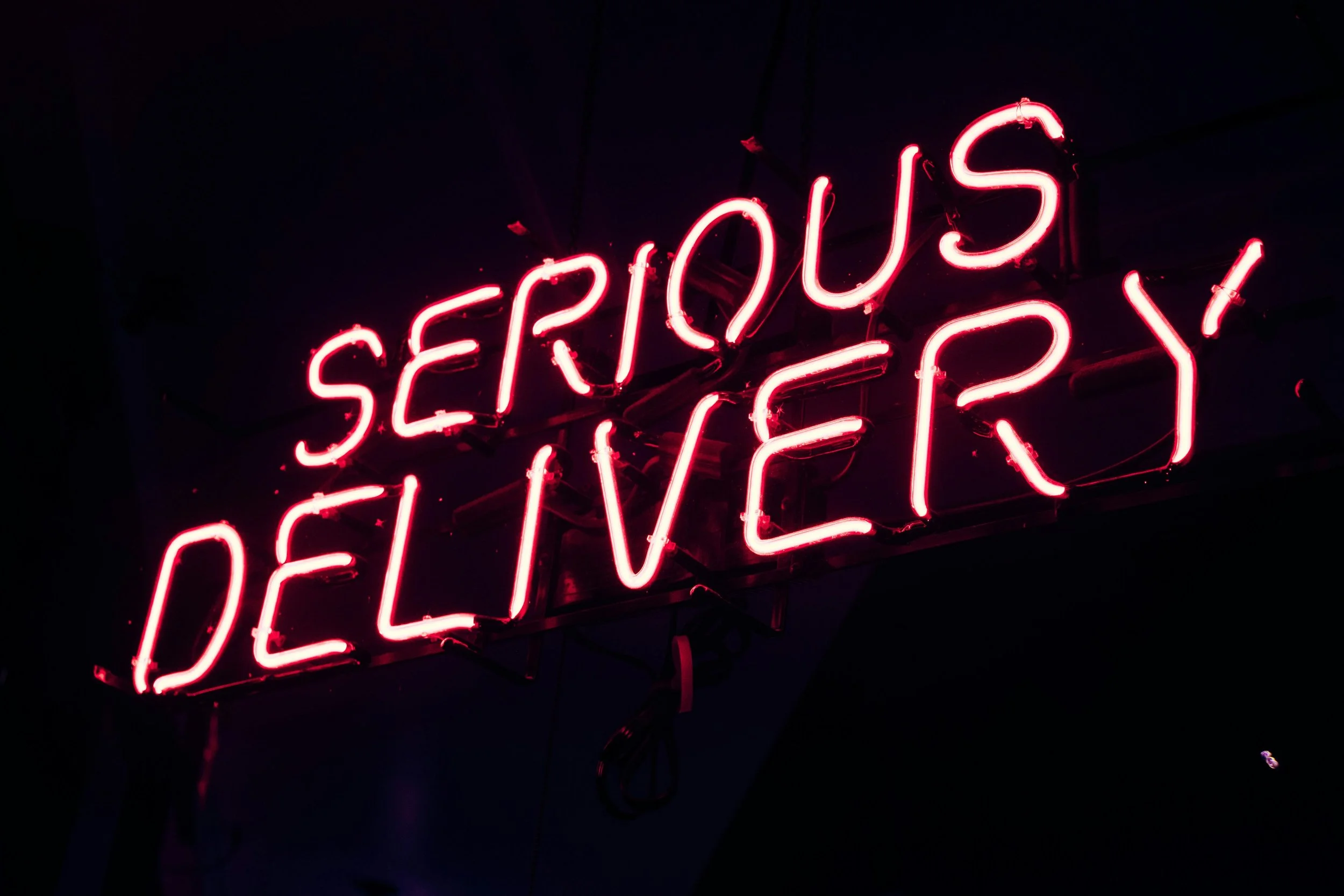 Neon sign reading 'Serious Deliver' in pink, mounted on a dark wall.