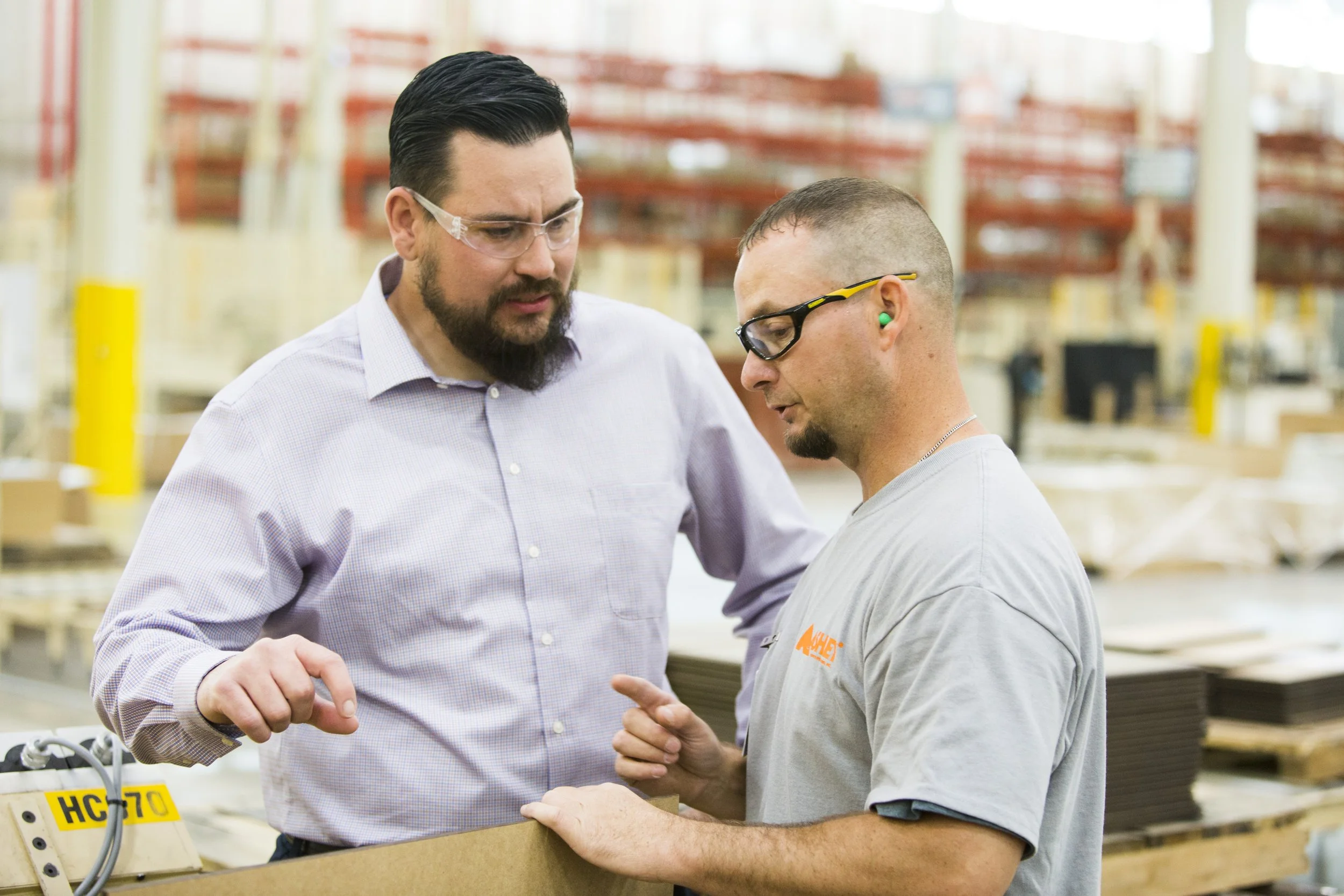 Two men in safety glasses and casual workwear discussing in a warehouse.