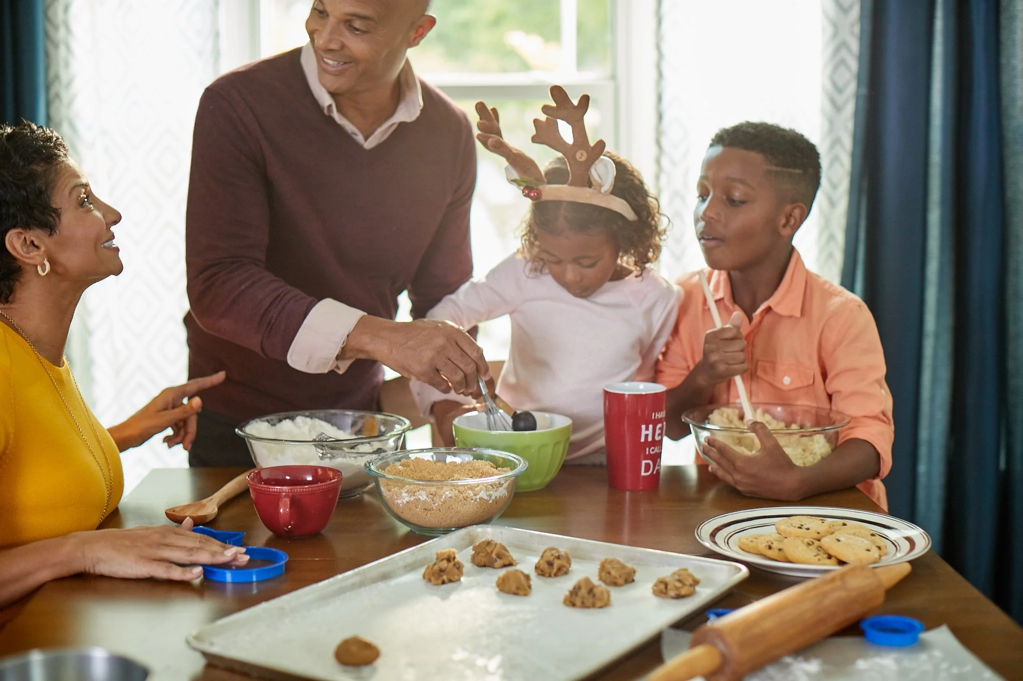 A family of four making cookies together in a bright kitchen, with a tray of cookie dough and baking tools on the table.
