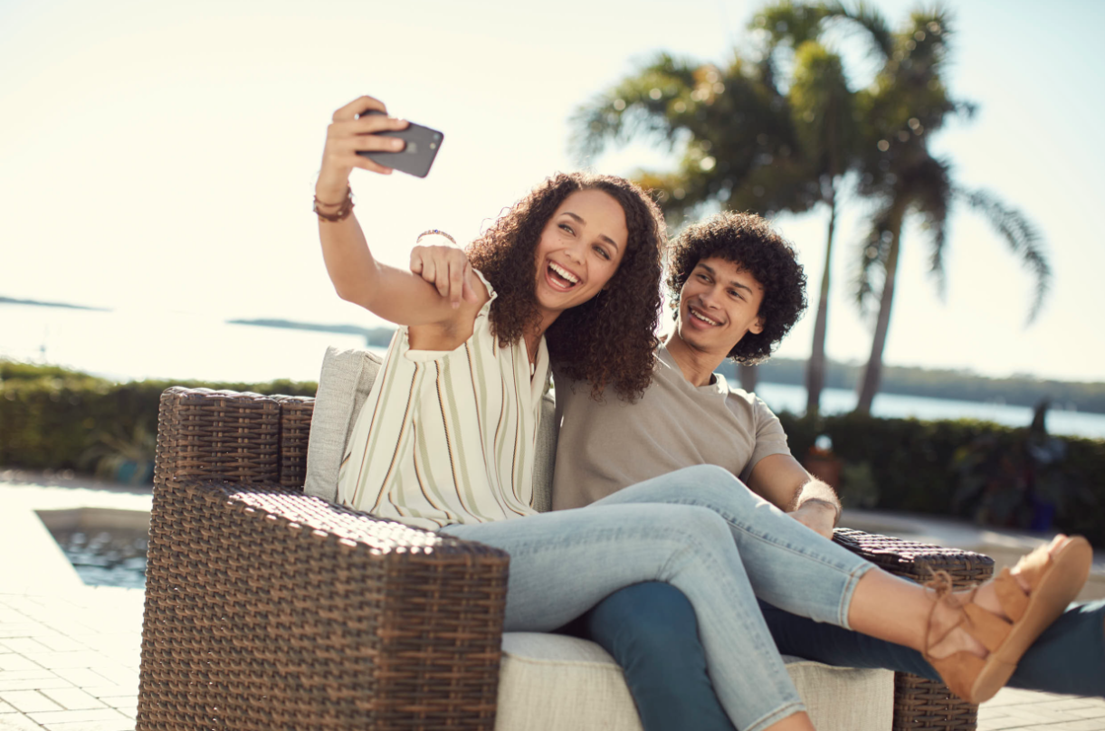 A young woman and man sitting on a wicker outdoor sofa, taking a selfie with a smartphone, smiling and enjoying a sunny day near the water with palm trees in the background.