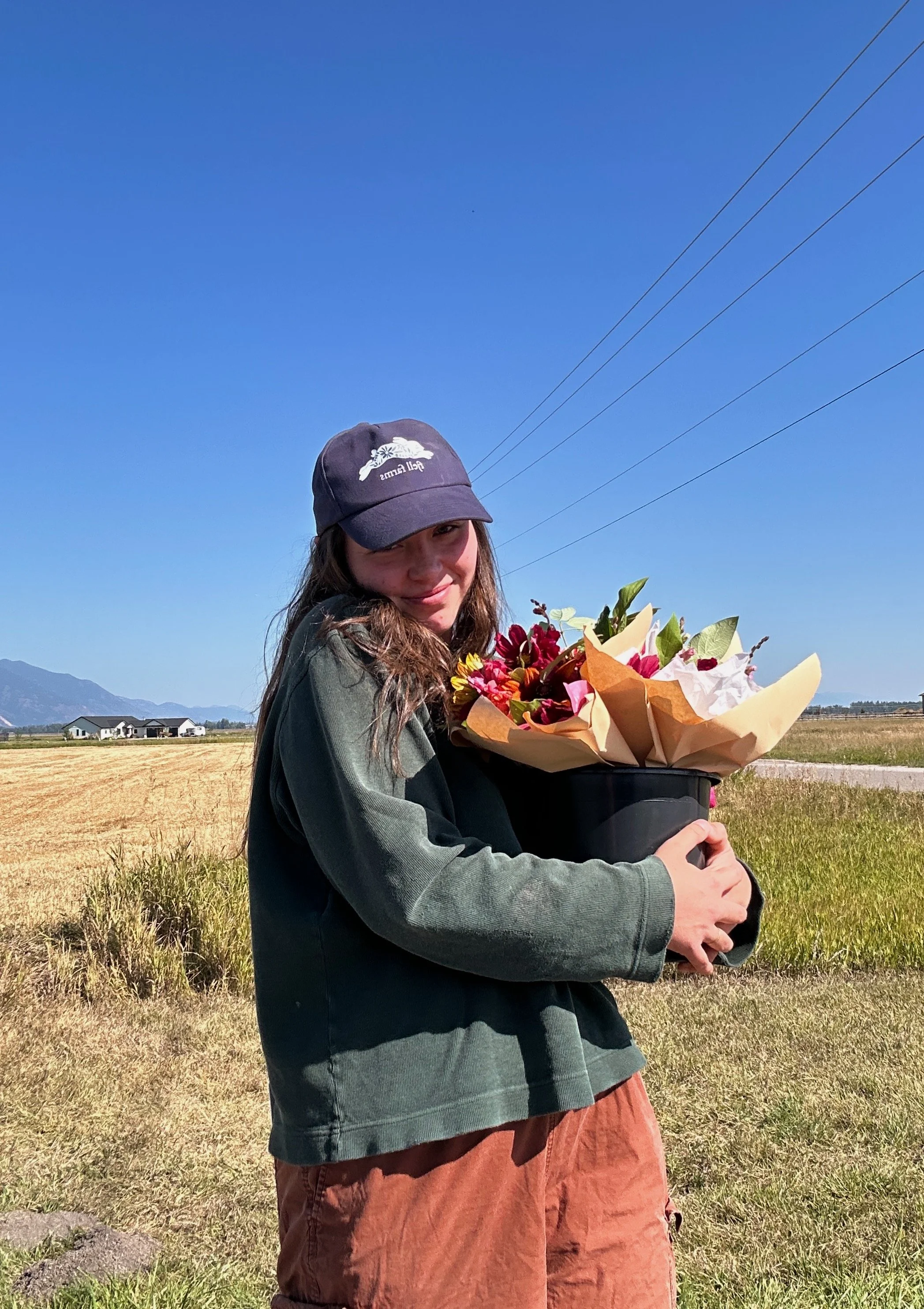 A young girl smiling and holding a bouquet of flowers outdoors on a sunny day, standing in a rural area with fields and mountains in the background.