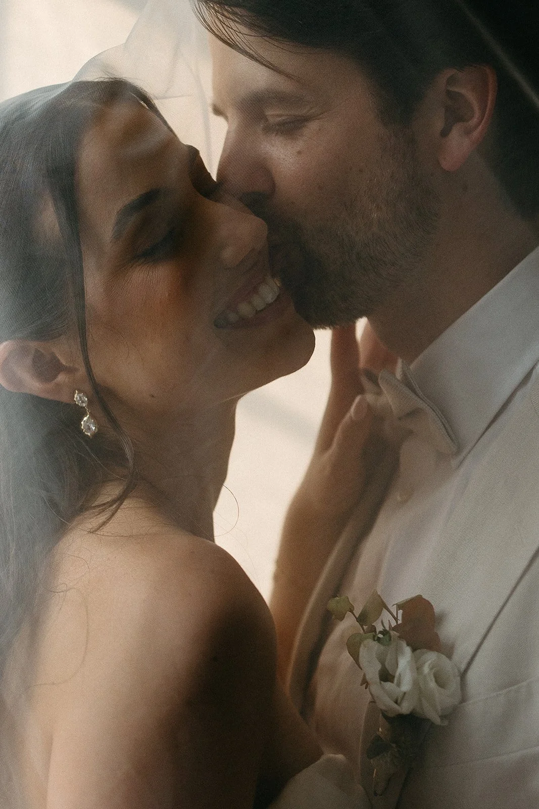 A close-up photograph of a joyful couple sharing a kiss, with the woman smiling and dressed in earrings and a strapless dress, and the man wearing a white shirt with a boutonniere.