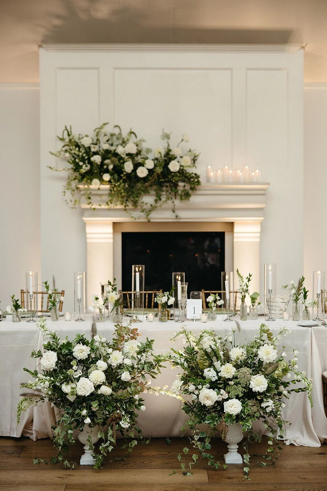 Elegant wedding reception table with white floral arrangements, candles, and gold chairs in front of a fireplace with a large floral display and candles on the mantle.