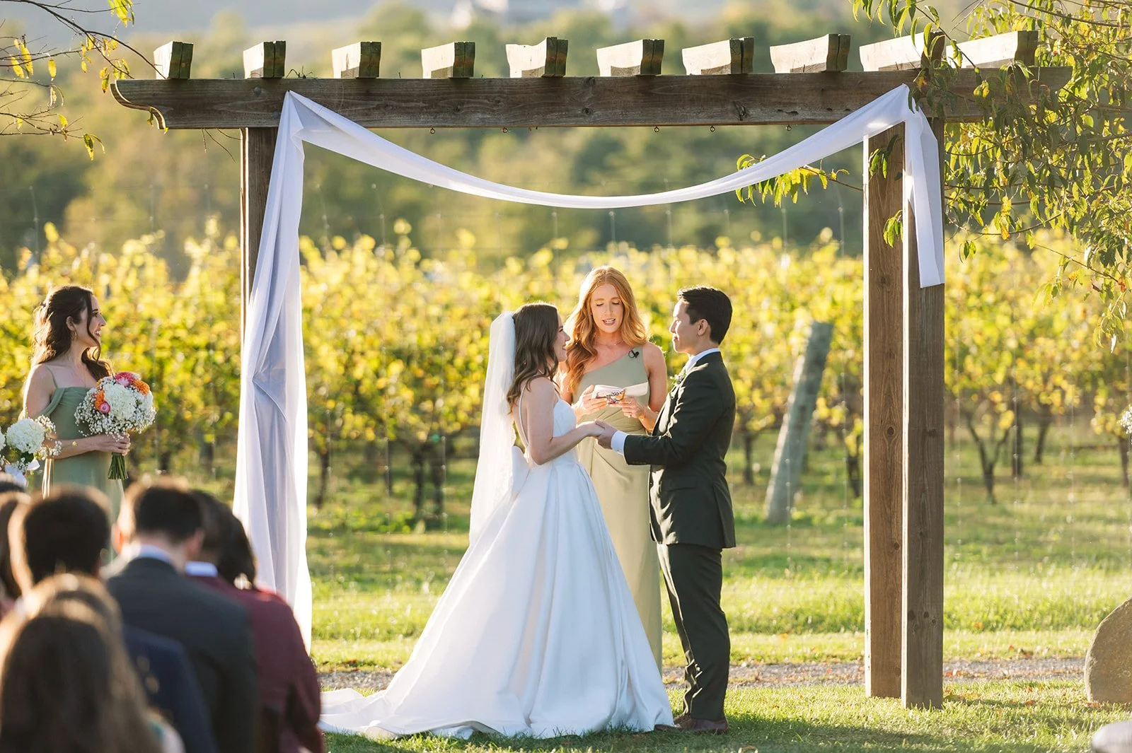 A couple getting married outdoors under a wooden arch decorated with white fabric, with a woman officiant reading from a booklet. The bride is in a white wedding gown, and the groom is in a black suit. Guests seated on the lawn watch the ceremony, with a scenic vineyard background.