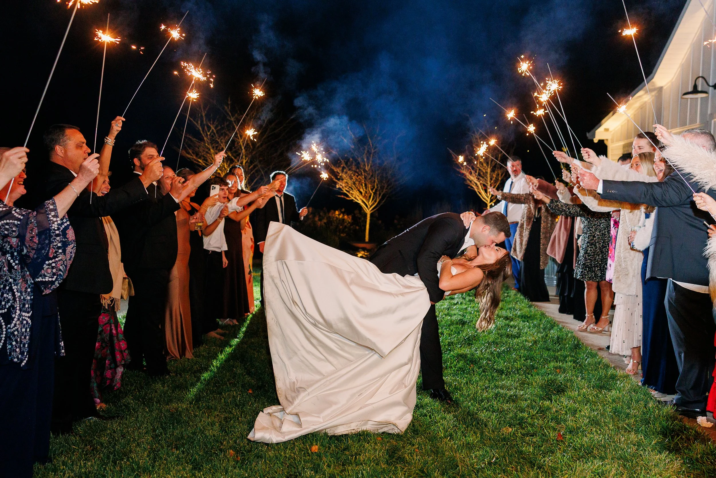 A newlywed couple sharing a kiss while being dipped by the groom during a nighttime celebration, surrounded by guests holding sparklers.