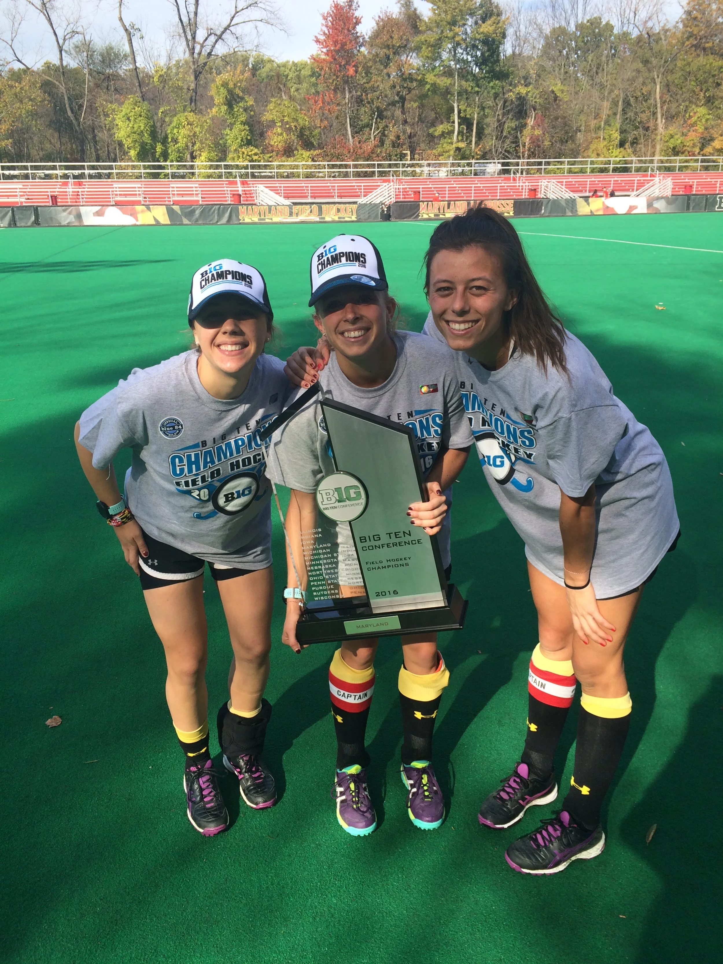 Three young female field hockey players celebrating on a green field with a tournament trophy. All three are smiling and wearing gray T-shirts, black shorts, and black knee-high socks with yellow and red that say 'Captain.' Two of them are wearing caps that say 'Big Champions'.