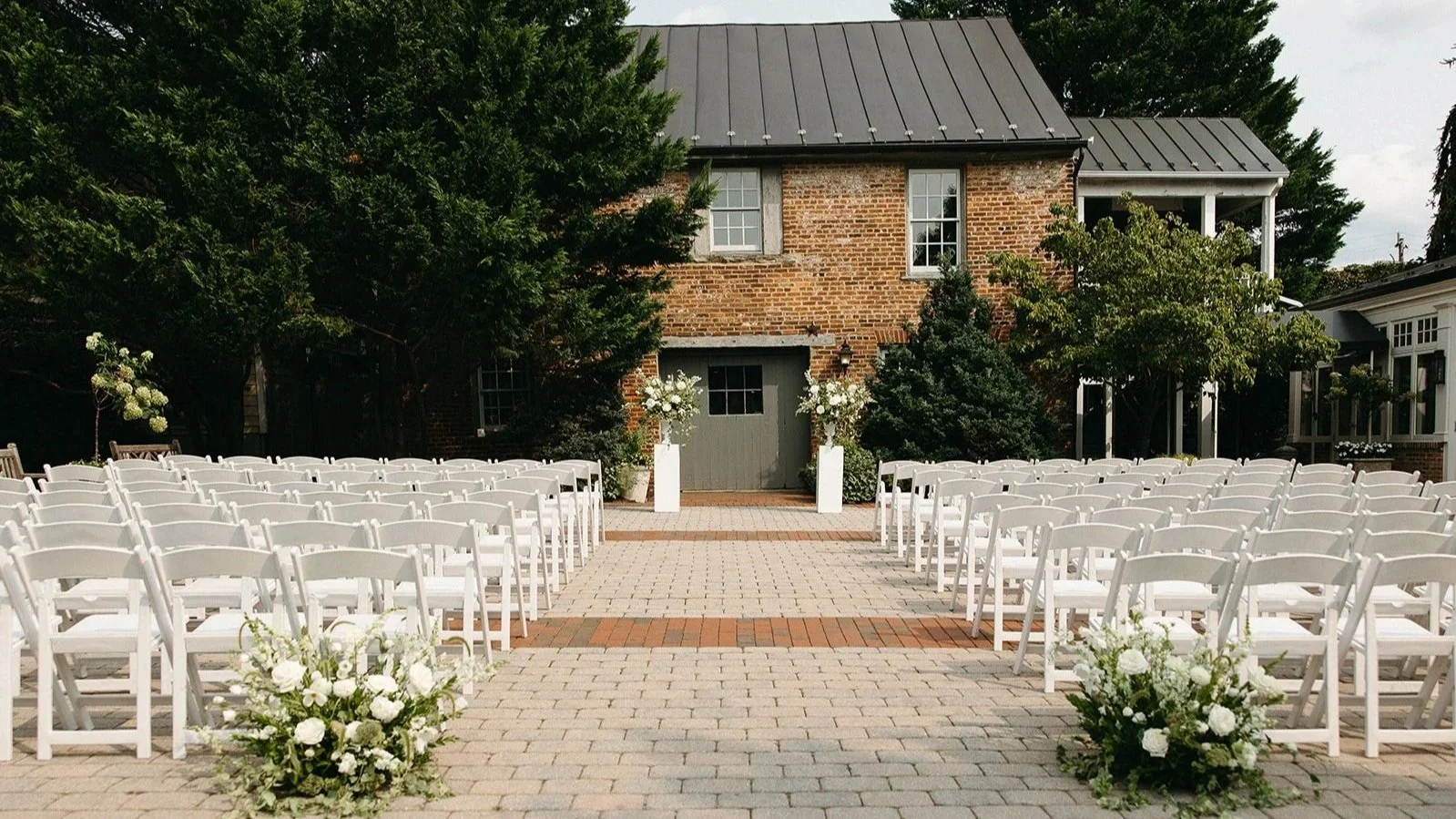 Outdoor wedding ceremony setup with rows of white folding chairs facing an altar with floral arrangements, in front of a brick building with trees and a cloudy sky in the background.