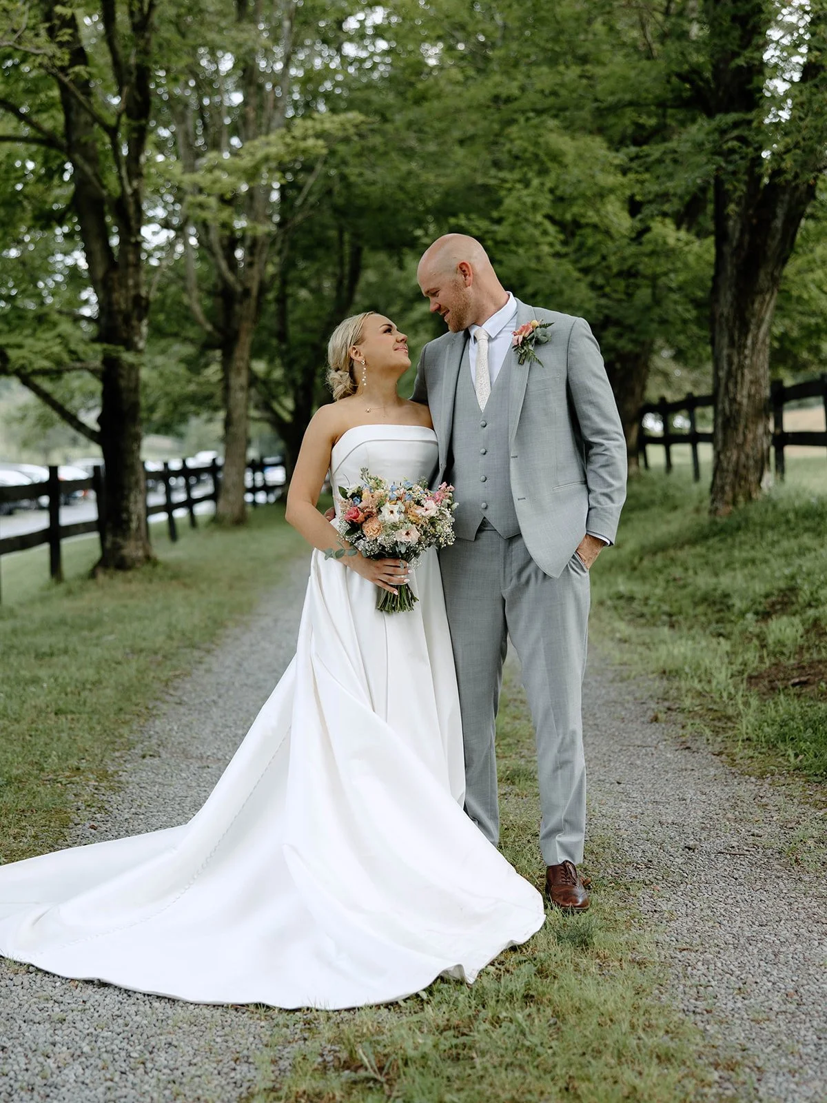 Bride and groom standing on a gravel path in a lush green outdoor setting, smiling at each other, with the bride holding a bouquet of flowers.