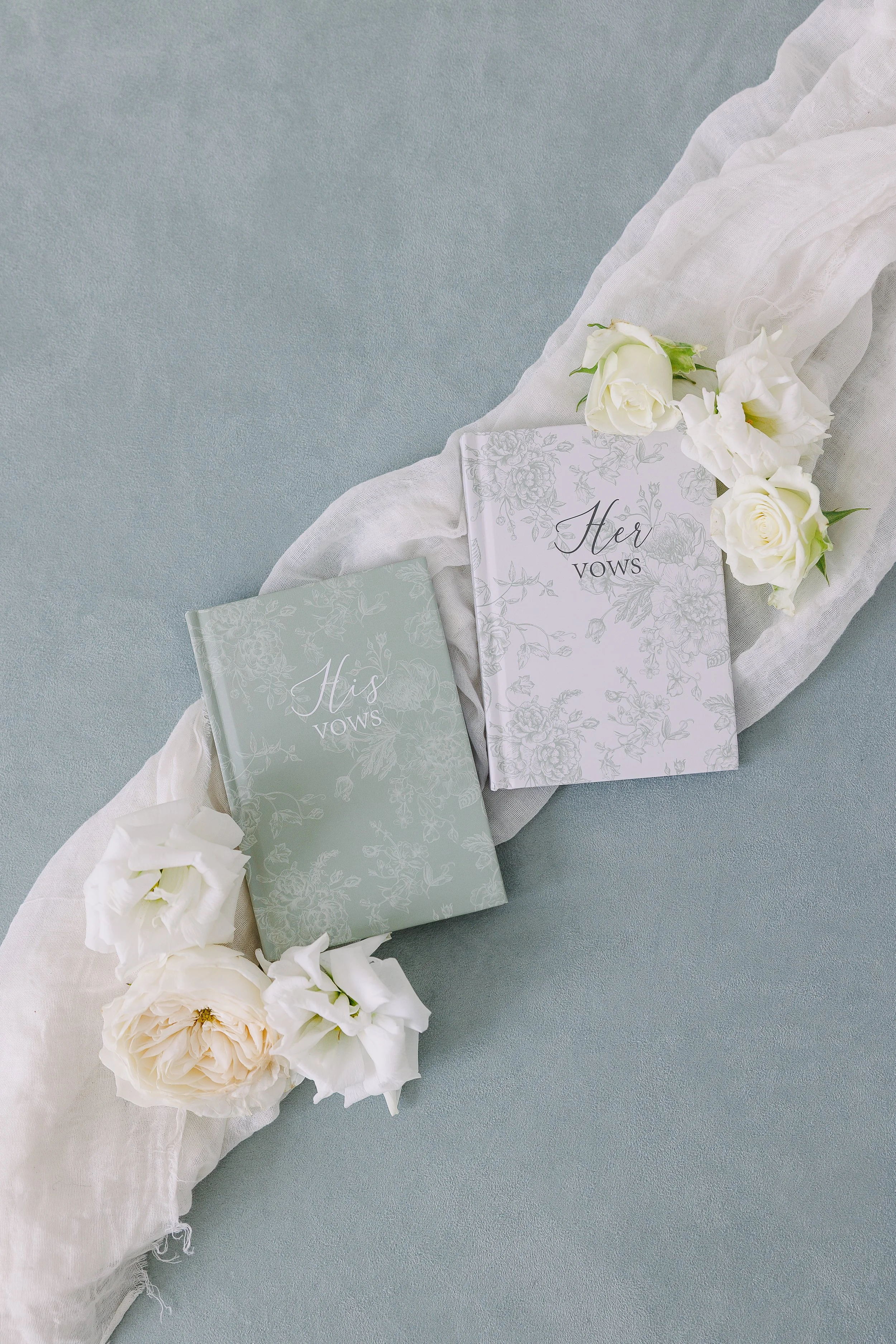 Wedding vow books labeled 'His Vows' and 'Her Vows' with white roses and a white cloth on a light teal background.