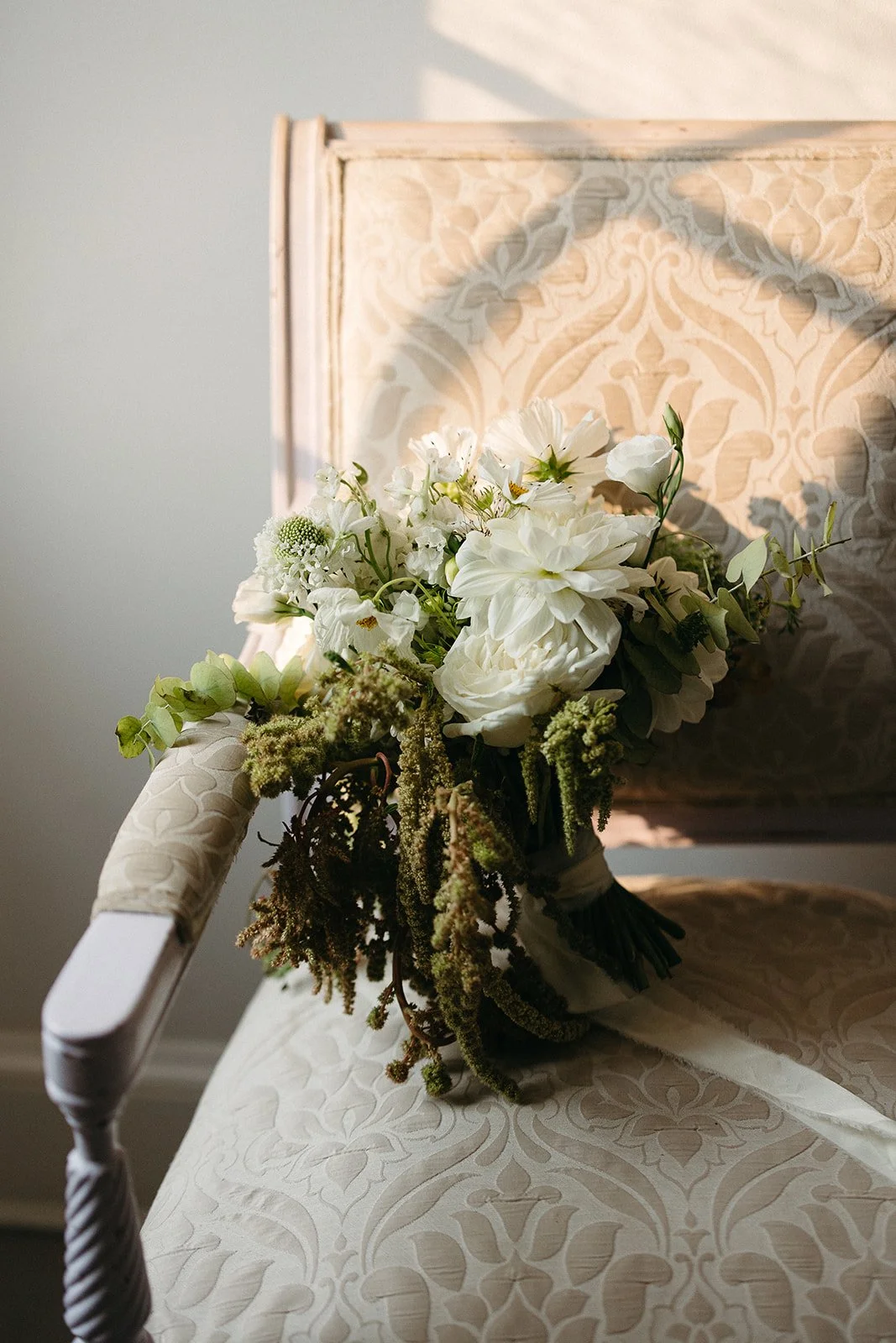 A bouquet of white flowers resting on an elegant patterned upholstered chair with sunlight casting shadows on it.