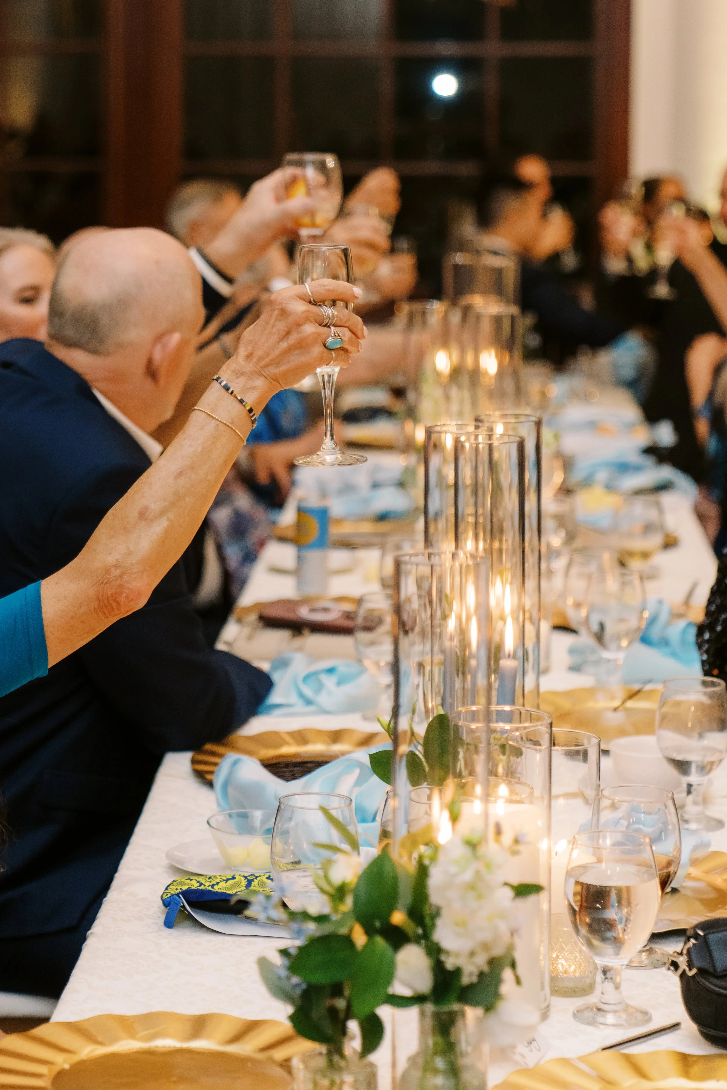 People raising glasses at a banquet table with gold plates and blue napkins, decorated with candles and floral arrangements.