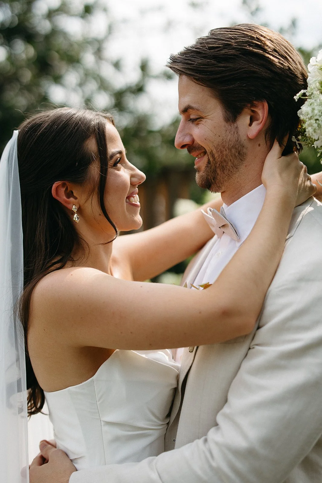 A bride and groom gazing at each other and smiling, with arms around each other, outdoors with greenery and white flowers in the background.