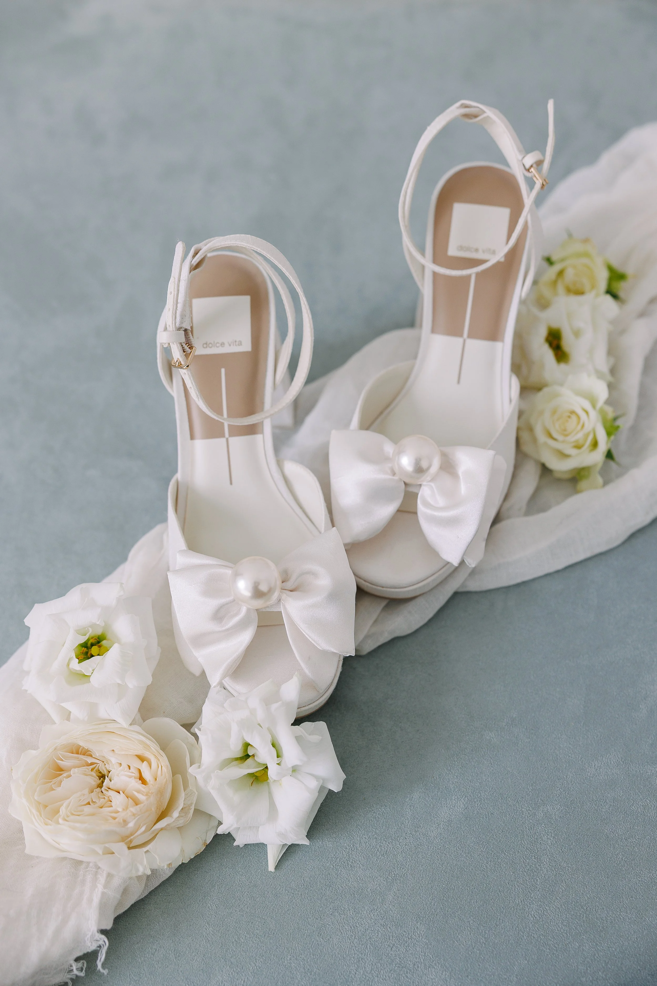 A pair of white high-heeled shoes with ankle straps, each adorned with a large satin bow with a pearl at the center, placed on a soft surface surrounded by white roses and flower petals.