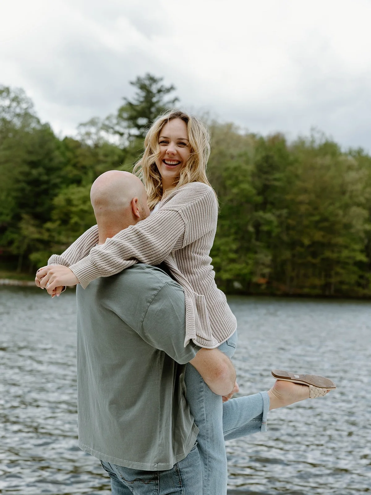 A woman with blonde hair and a beige sweater smiling while being carried by a man near a lake with trees in the background.
