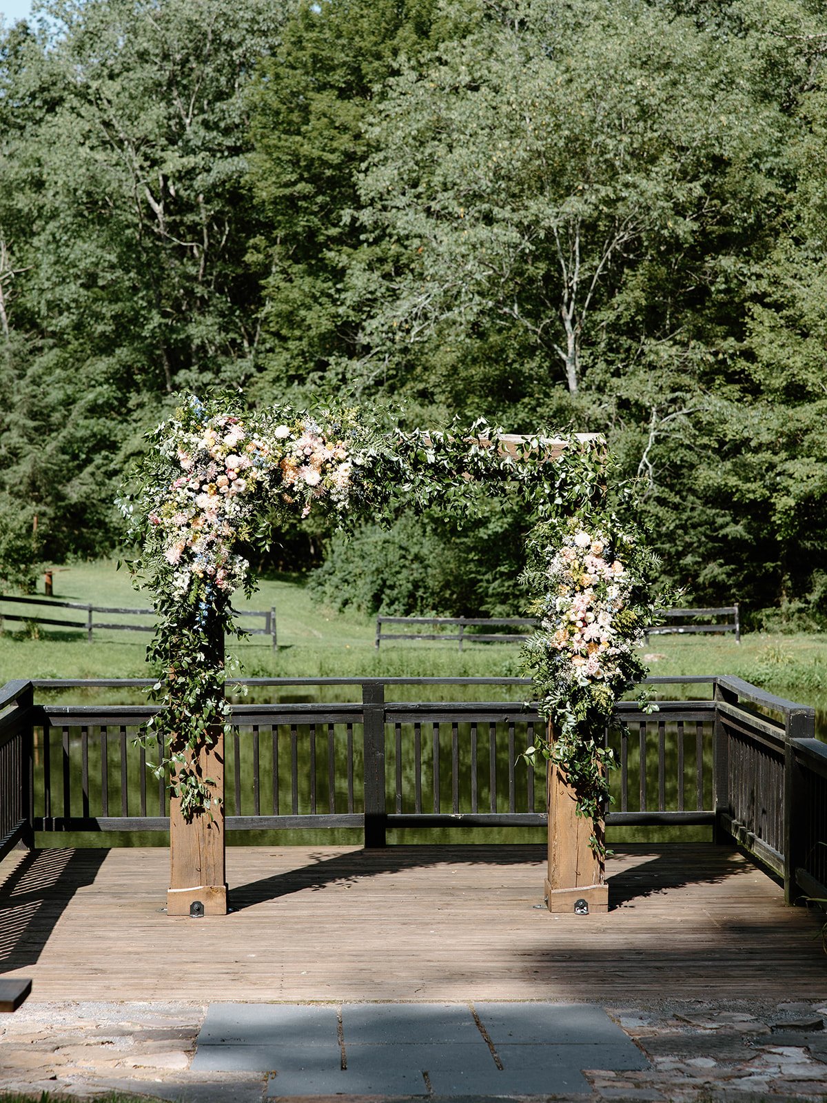 Wooden wedding arch decorated with white and pink flowers and greenery, set up outdoors on a wooden deck with trees and a grassy area in the background.