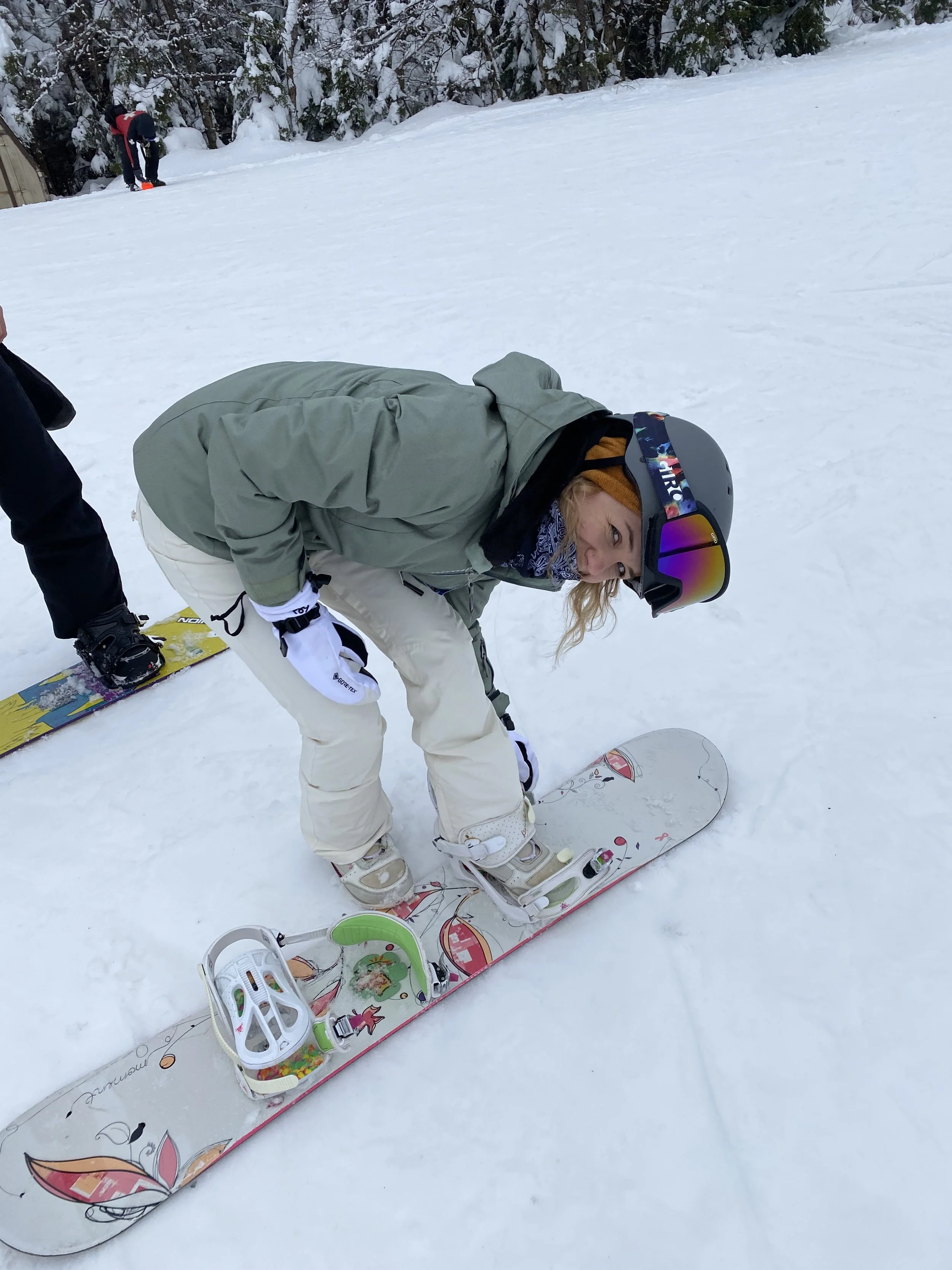 Woman in ski gear bending over on a snowboard on a snowy slope with trees in the background, wearing a helmet, goggles, and a scarf.