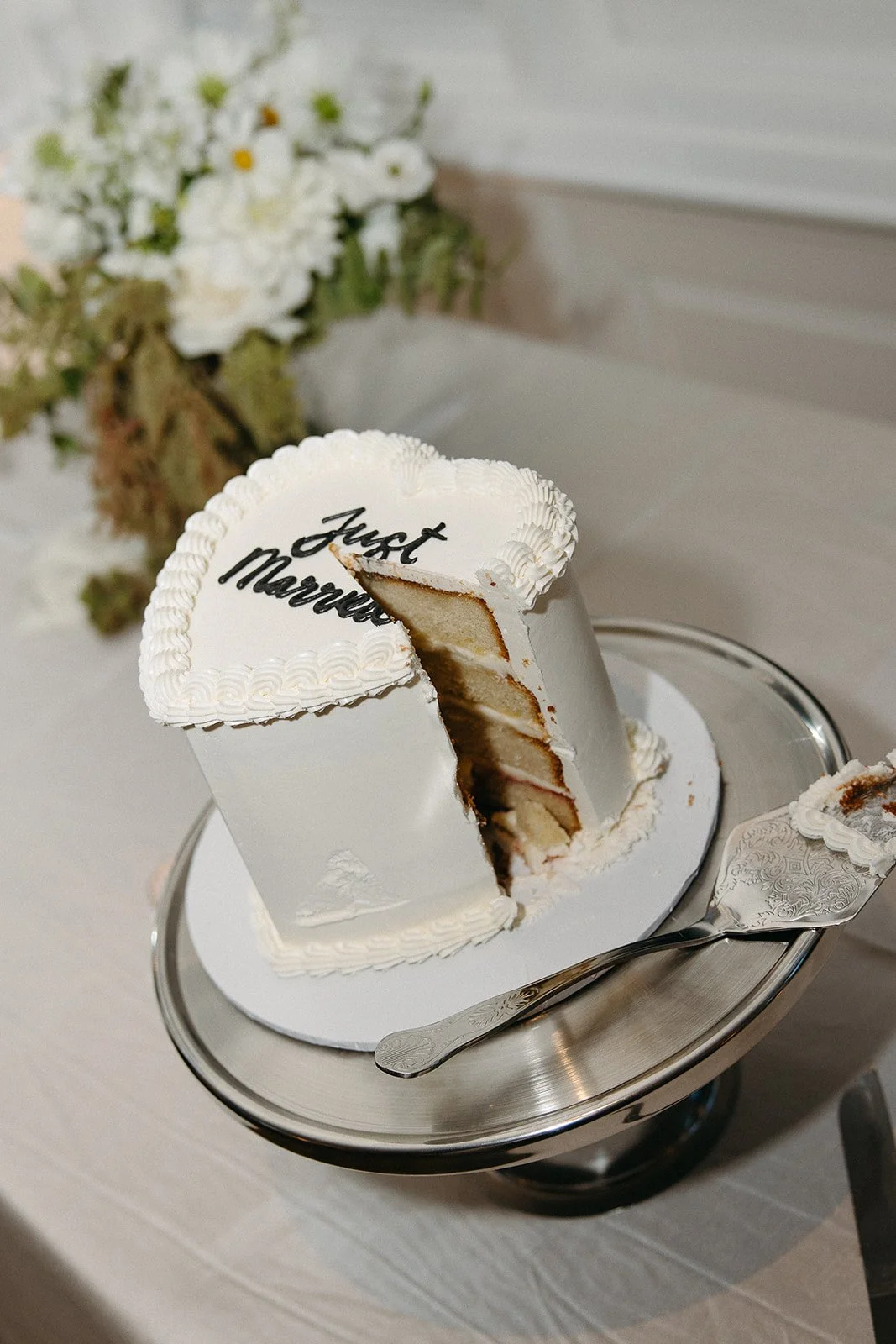 A partially cut white wedding cake with layers, decorated with white frosting and black cursive writing that says 'Just Married', placed on a silver cake stand with a cake server beside it, and a bouquet of flowers in the background.